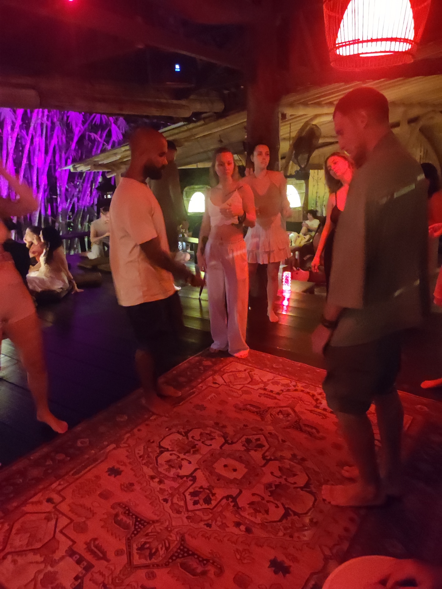 The image captures an indoor gathering, possibly a party or social event, illuminated by a red light source, perhaps a decorative lamp or stage lighting. The setting appears to be a bar or lounge in Lokaserena, Indonesia with wooden structures, some wrapped with purple foliage or lights.
Several individuals are present. In the foreground, a bald man with a beard is wearing a light-colored t-shirt and dark shorts. Next to him are two women in light colored outfits. Other individuals are seated or standing around the room, engaged in conversation or observation. A large, patterned rug covers the floor in the foreground.