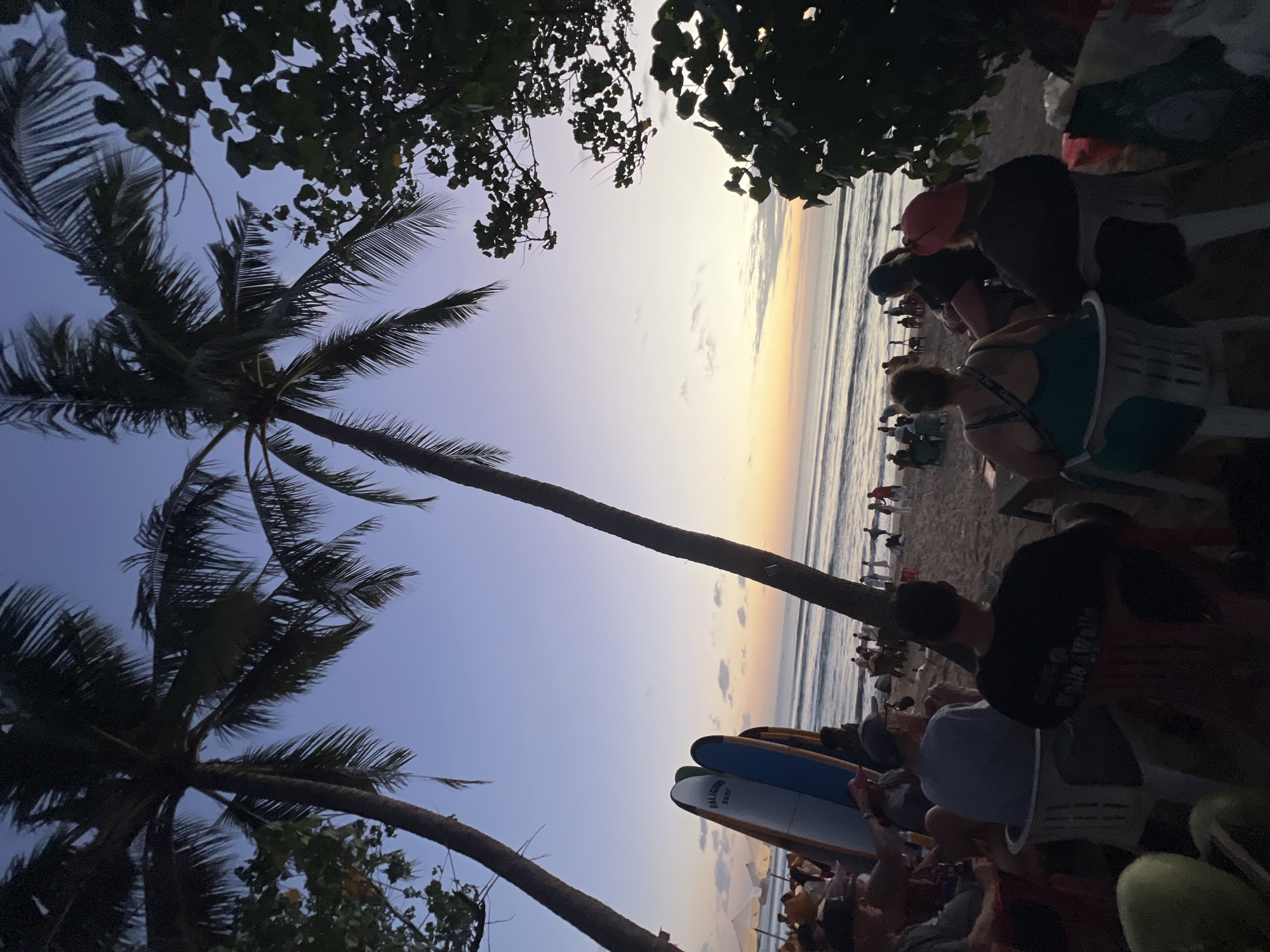 This is a vertical, eye-level, full-shot photograph of a beach scene at dusk in Kuta, Indonesia. Two palm trees frame the shot, with their fronds silhouetted against a gradient sky that transitions from pale blue at the top to soft orange and yellow at the horizon. The ocean is calm with gentle waves, and a few small boats are visible in the distance. On the beach, several people are seated on chairs, facing the ocean and observing the sunset. Three surfboards stand upright in the sand near the seated individuals. Further down the beach, other people are scattered about, some standing and interacting, while others appear to be walking. The overall atmosphere is relaxed and serene, indicative of a popular tourist spot at the end of the day.