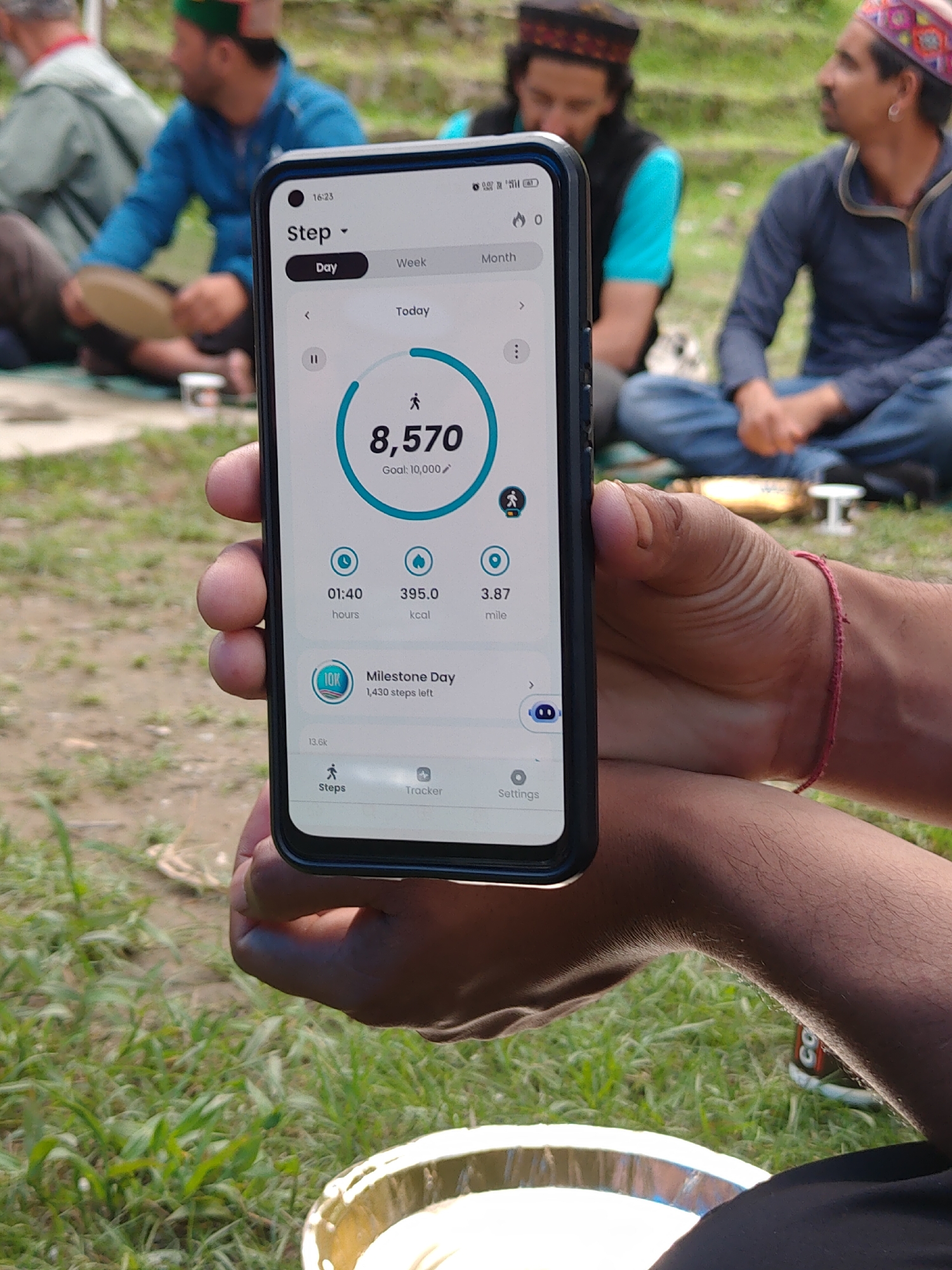 A person holds a smartphone displaying a step-tracking app outdoors. The phone screen shows 8,570 steps achieved today, with a goal of 10,000 steps. The app also displays calories burned and distance covered in miles. In the background, two individuals are seated on the ground, with one wearing a blue jacket and the other a dark hooded sweatshirt and a patterned cap. The environment appears to be a grassy outdoor area, likely in Majhach, India, suggested by the clothing and setting. The overall scene captures a moment of activity tracking amidst a relaxed outdoor gathering.