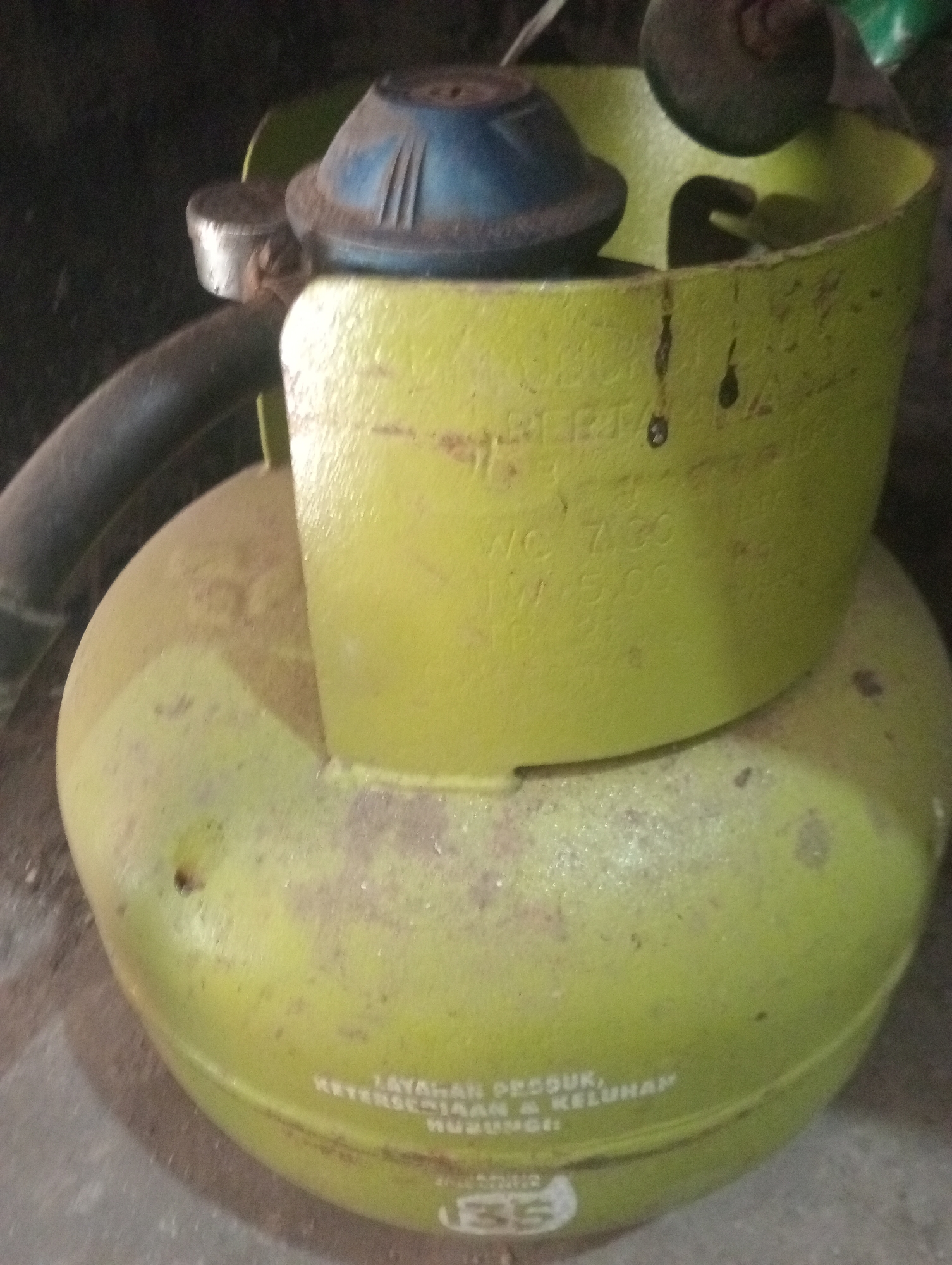 The image shows a close-up of a green gas cylinder with a blue regulator. The regulator has a metal clamp connecting it to the cylinder. There is writing on the cylinder that appears to be in Indonesian. The cylinder is sitting on a surface that looks like concrete or stone. The background is dark and out of focus. There are no people or animals visible in the image. The scene is indoors or in a shaded outdoor area. The location in Buduan, Indonesia, is not visually discernible from the image itself.