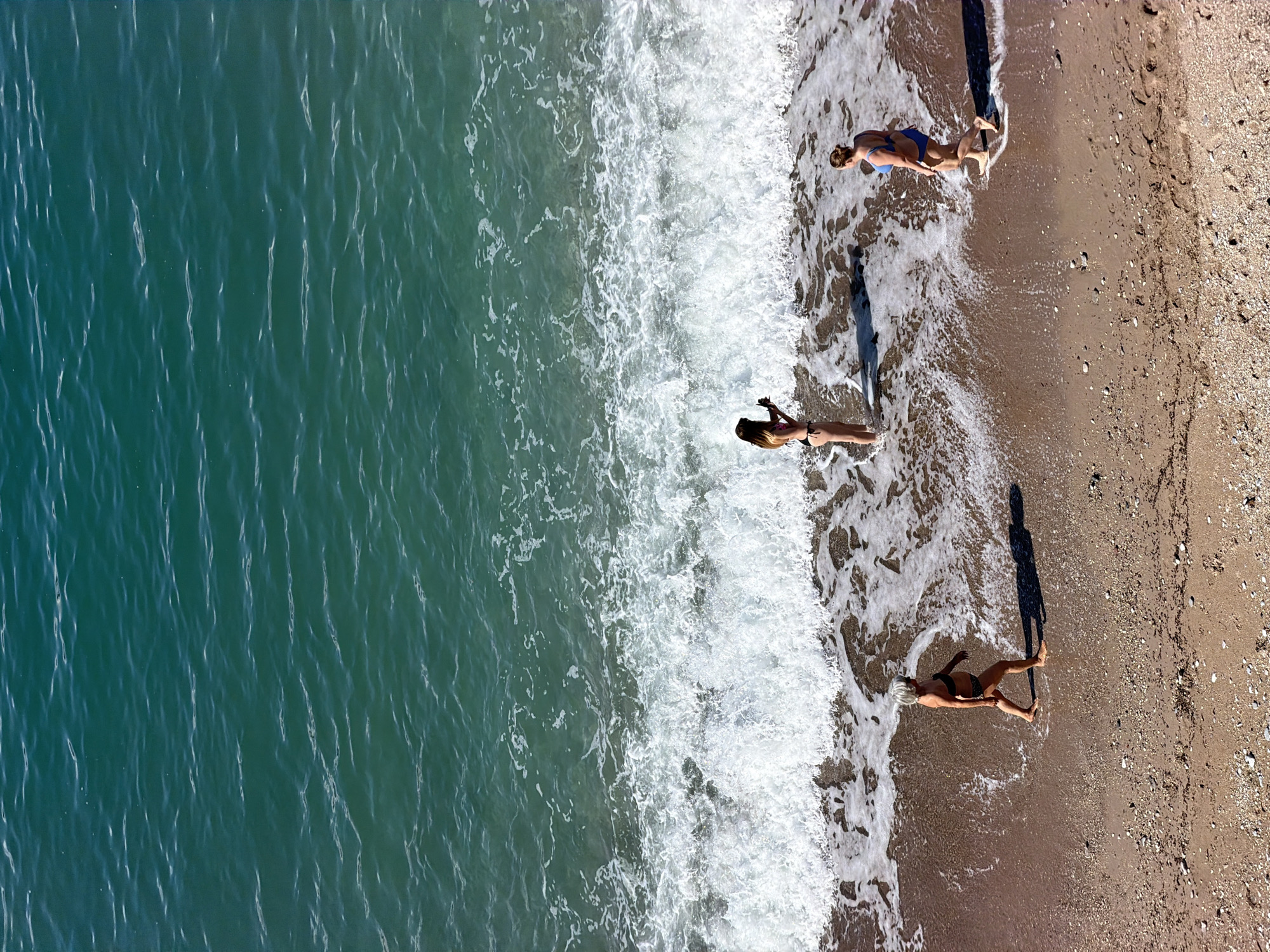 This aerial, top-down image presents a vibrant coastal scene at Boreti, Montenegro, featuring a clear turquoise-green sea meeting a sandy, pebbled beach. The image is divided diagonally, with the ocean occupying the left side and the beach on the right. A distinct line of white, foamy waves marks the shoreline where the water actively washes onto the land.
Three individuals, all appearing to be female, are visible on the wet sand, near the water's edge. From bottom to top along the image's right side, the first person is lying prone on their stomach, wearing a dark bikini with a patterned bottom. Slightly above, a second person stands upright, facing the ocean, also in a dark bikini, seemingly holding something in their hands. The third person, positioned furthest up the beach near the water, stands in a blue one-piece swimsuit.
A notable detail is the presence of long, distinct shadows cast by each individual across the wet sand, extending towards the ocean. These elongated shadows strongly suggest that the photograph was taken either in the early morning or late afternoon, when the sun is low in the sky. The bright lighting indicates clear, sunny weather. The overall scene depicts a leisurely moment by the sea. There is no visible text in the image.