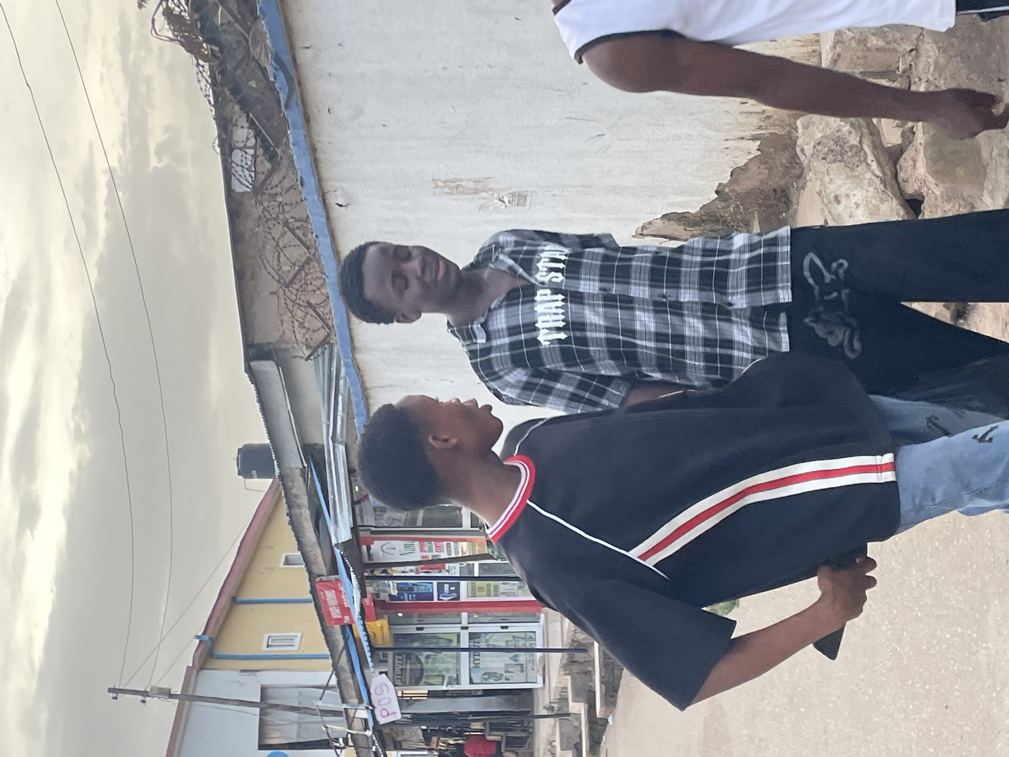 Two young men are standing on a sidewalk in Jos, Nigeria. One is wearing a black shirt with red and white stripes and blue jeans, and holding a phone in his hand. The other is wearing a black and white checkered shirt with the words "TRAP STAR" printed on it, and black pants. Behind them is a row of storefronts, and a building with a yellow roof and blue trim. The sky is overcast. There is barbed wire strung along the top of a nearby wall. A third person, only partially visible, is standing near the two young men, wearing a white shirt. The men are facing each other. The location appears to be an urban area, likely a commercial district.