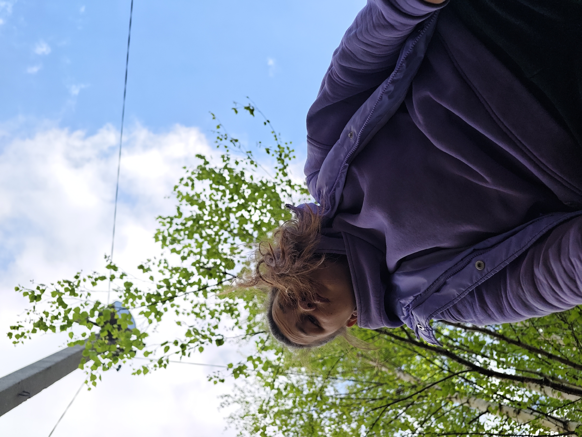A low-angle shot shows a person wearing a purple hooded jacket standing in an outdoor environment. The individual's face is visible, with light brown hair. The background consists of trees with green leaves against a partly cloudy blue sky. A concrete pillar or post is visible on the left side of the frame, near the trees. Given the stated location of Chaikovsky, Russia, the trees are likely species common to that region. The photograph's orientation suggests the subject is looking down at the camera.