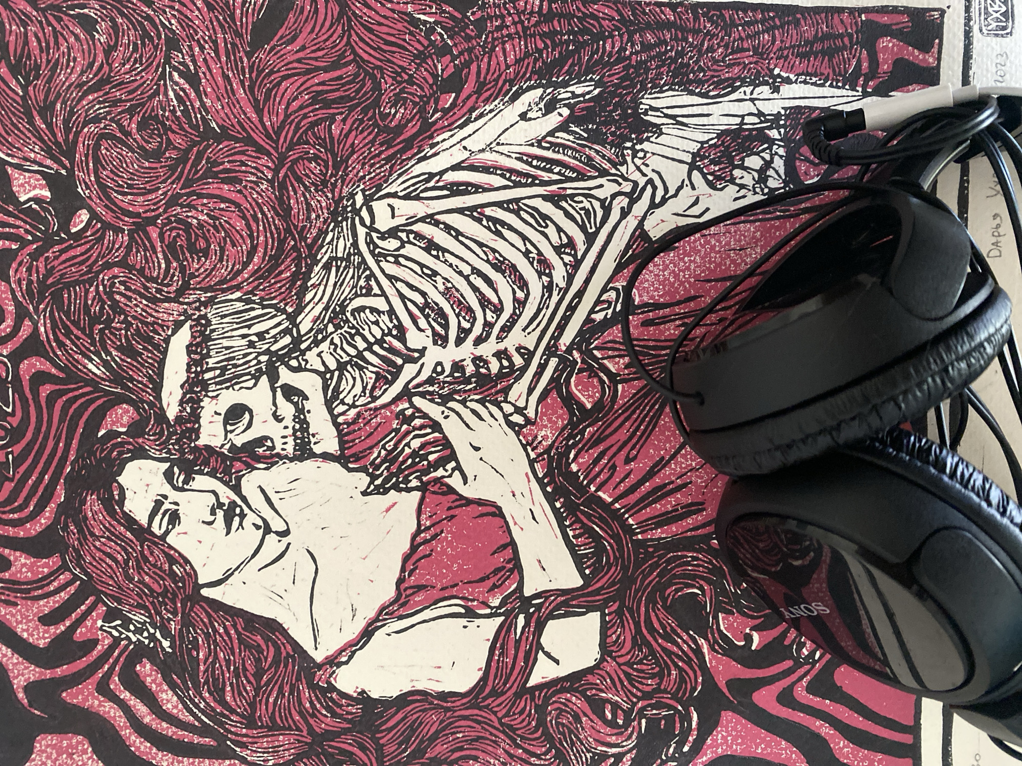 The image shows a close-up of a print depicting a woman lying beneath a skeleton. The artwork is rendered in a dark pink and black color scheme with swirling patterns forming hair or decorative elements. A pair of black Sony headphones with a visible cable are positioned on top of the artwork, partially obscuring the image. In the upper right corner, there's a small section of paper with some handwritten text including the number "2023." The location, Rusanovo, Russia, does not appear to be represented in the image.