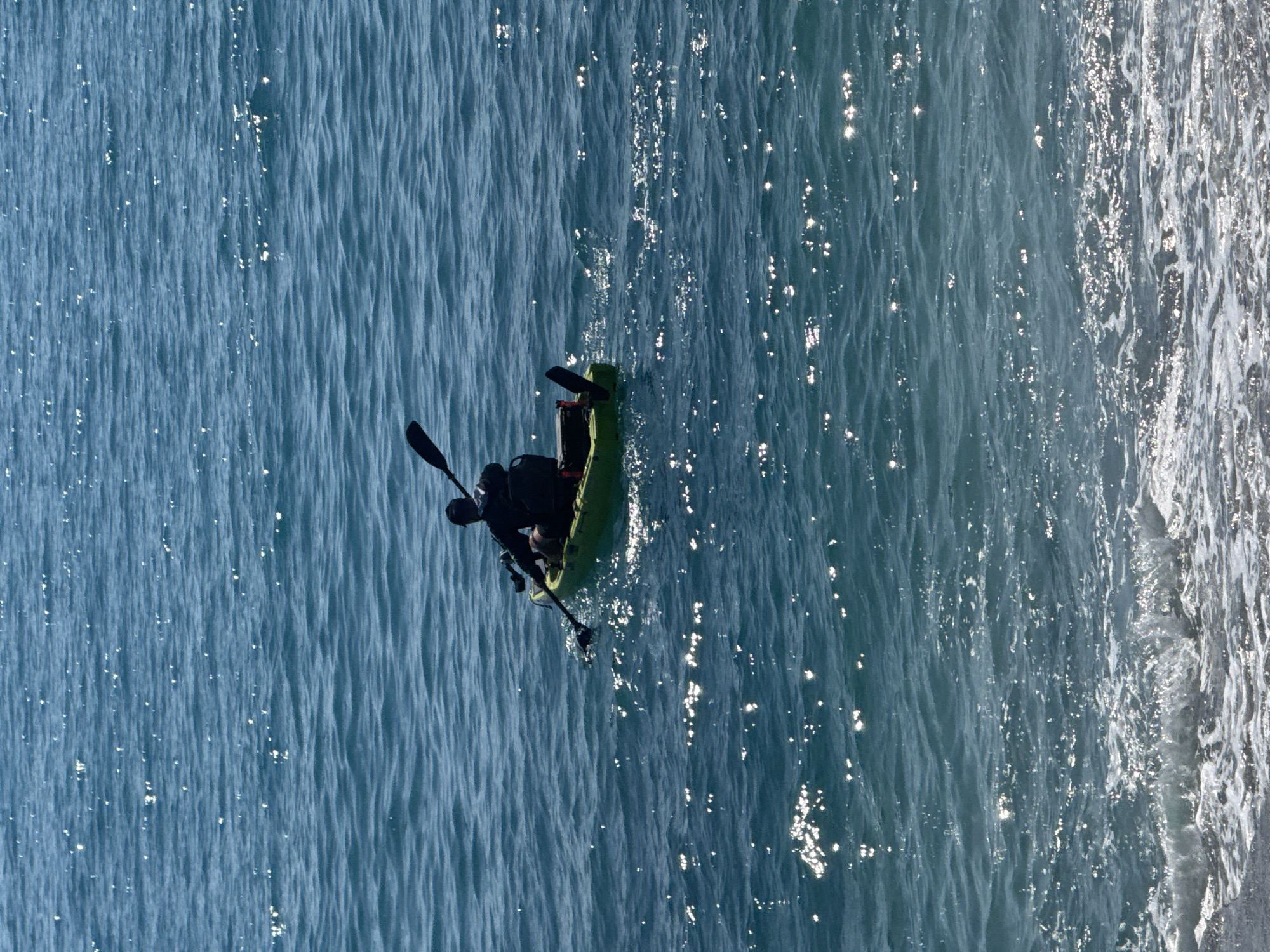 A person paddles a light green kayak in the ocean, near the shore. The ocean water appears blue with patches of sunlight reflecting on the surface, creating a shimmering effect. A set of paddles is located on both sides of the kayak. The individual in the kayak is wearing dark clothing. The location is Unknown, Unknown.