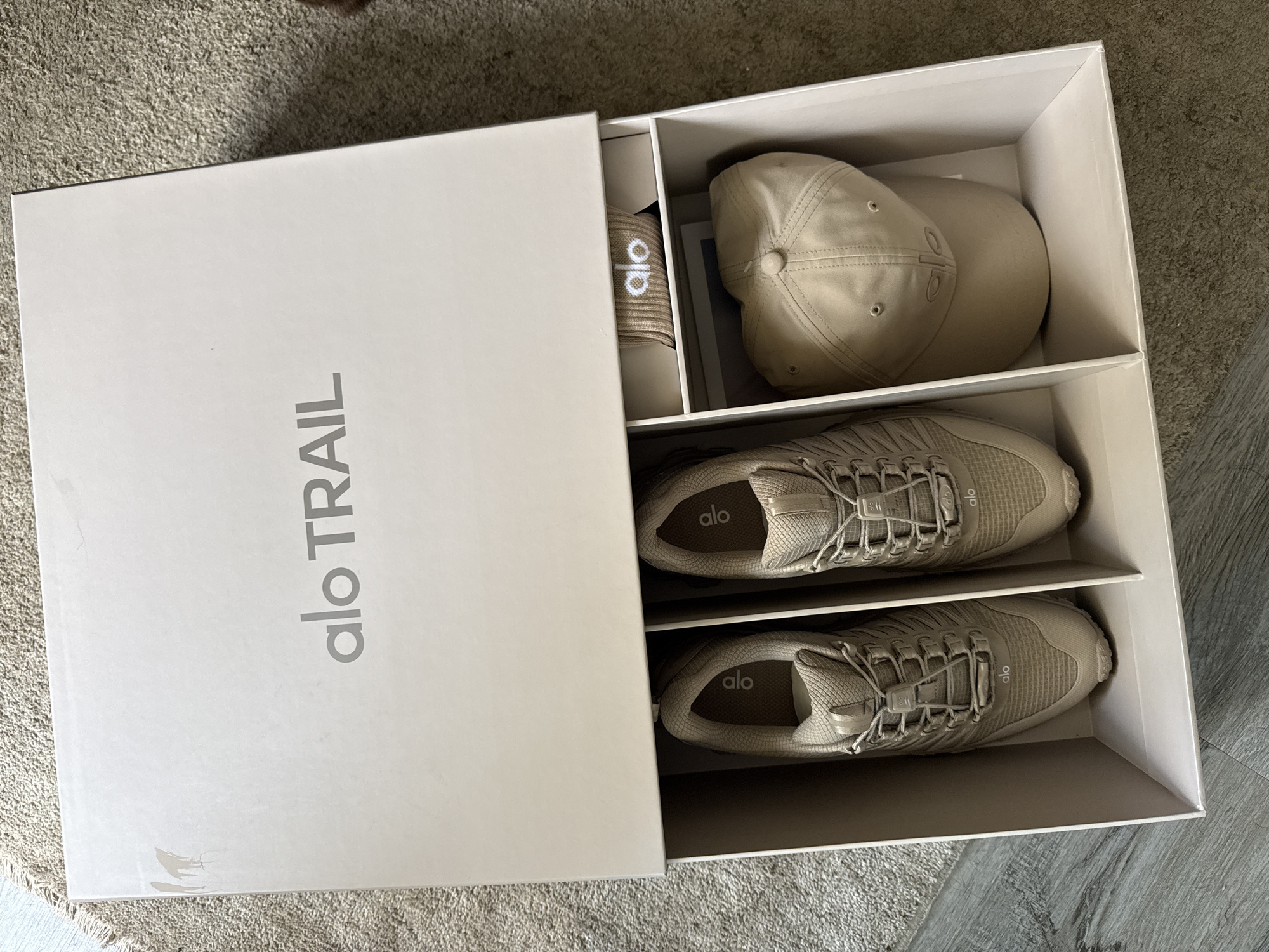 The image shows a white box labeled "Alo Trail" containing Alo brand merchandise. Inside the box, there are two pairs of Alo brand sneakers, a hat, and a headband. The items are all beige in color. The box is sitting on a carpeted floor. The location of the scene cannot be determined from this image.