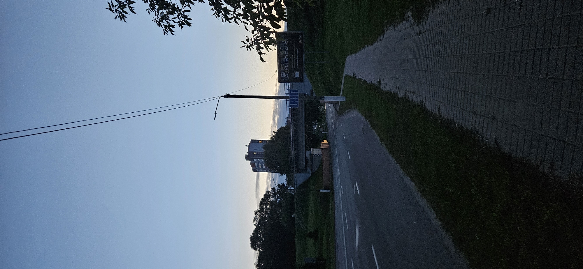 The image depicts a twilight scene on a street in Vitsebsk, Belarus, likely near an overpass. A paved road with white lane markings runs diagonally across the lower left of the frame, with a matching sidewalk to its right. Green grass borders both the road and the sidewalk. In the background, a multi-story building stands behind a bridge, which appears to be carrying a road over the one in the foreground. A street light pole with wires is visible to the right of the building. A billboard is positioned to the right of the street light pole. The sky is a pale blue, characteristic of dusk or dawn. No people are visible in the frame.