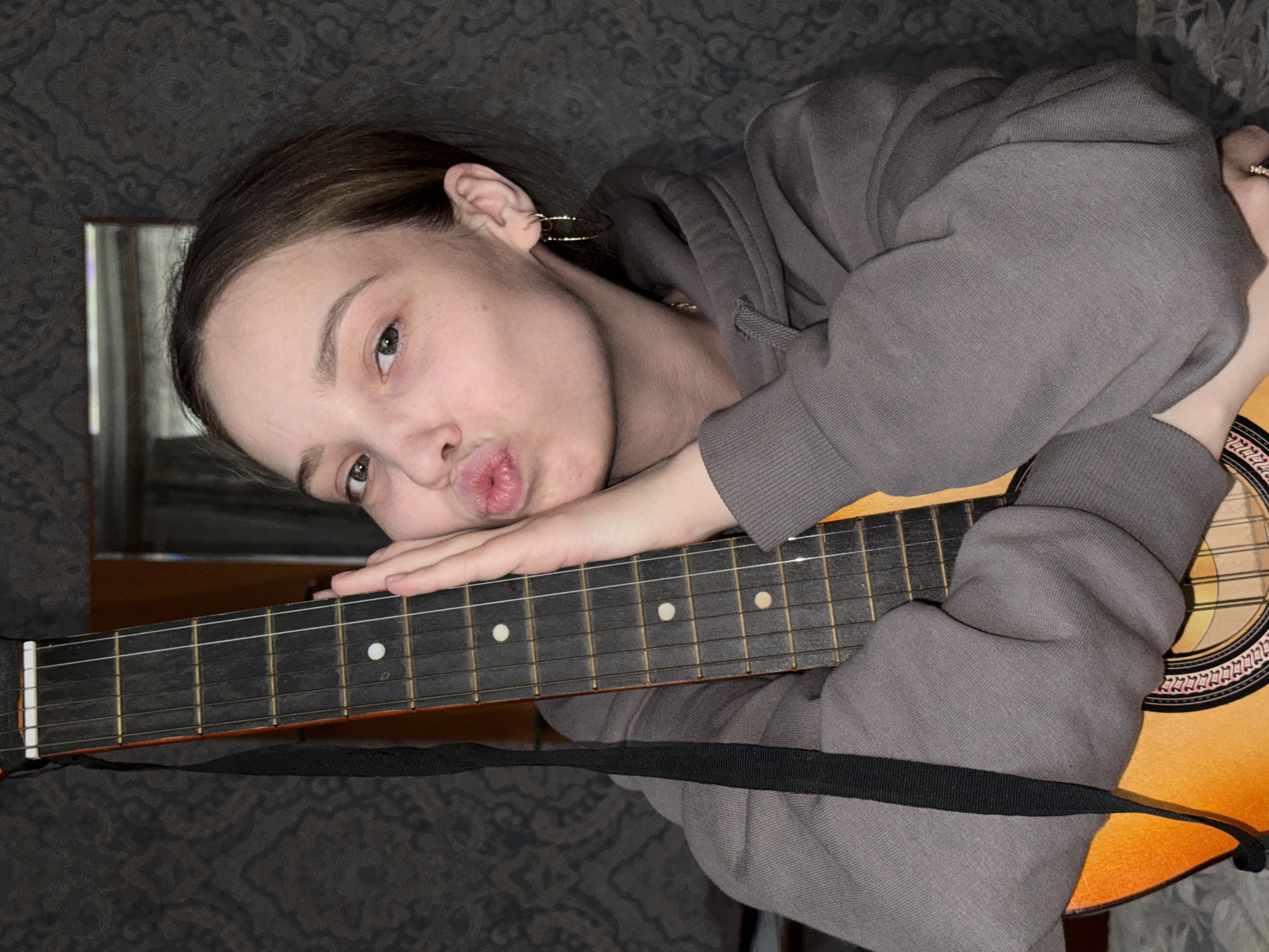 A young woman rests on a guitar in an indoor setting. She is light-skinned, with dark hair and wearing a grey hoodie. The guitar is an acoustic model with a dark fretboard and light-colored strings. A black strap is visible. The background features a patterned wall and a glimpse of what appears to be a mirror. The location is Прикамский, Russia.