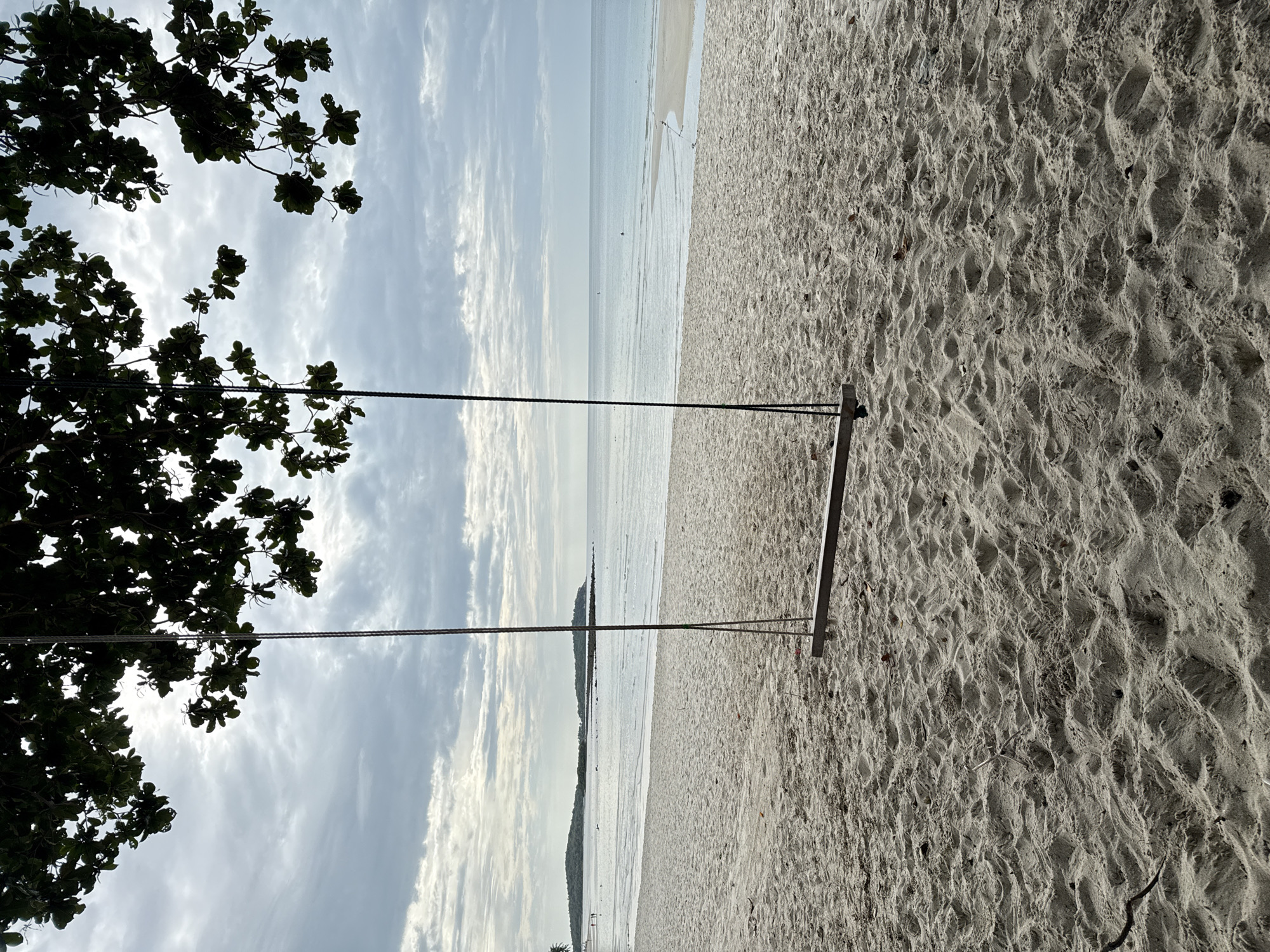 An empty swing hangs from the branches of a tree over a sandy beach on the island of Koh Samui, Thailand. The swing seat, made of wood, is suspended by two ropes. The foreground is dominated by rippled sand, with scattered dried leaves and twigs. In the midground, a calm ocean stretches to the horizon under a cloudy sky. Distant landforms, appearing as islands or peninsulas, are visible along the coastline to the left and right. The overall atmosphere is tranquil and serene.