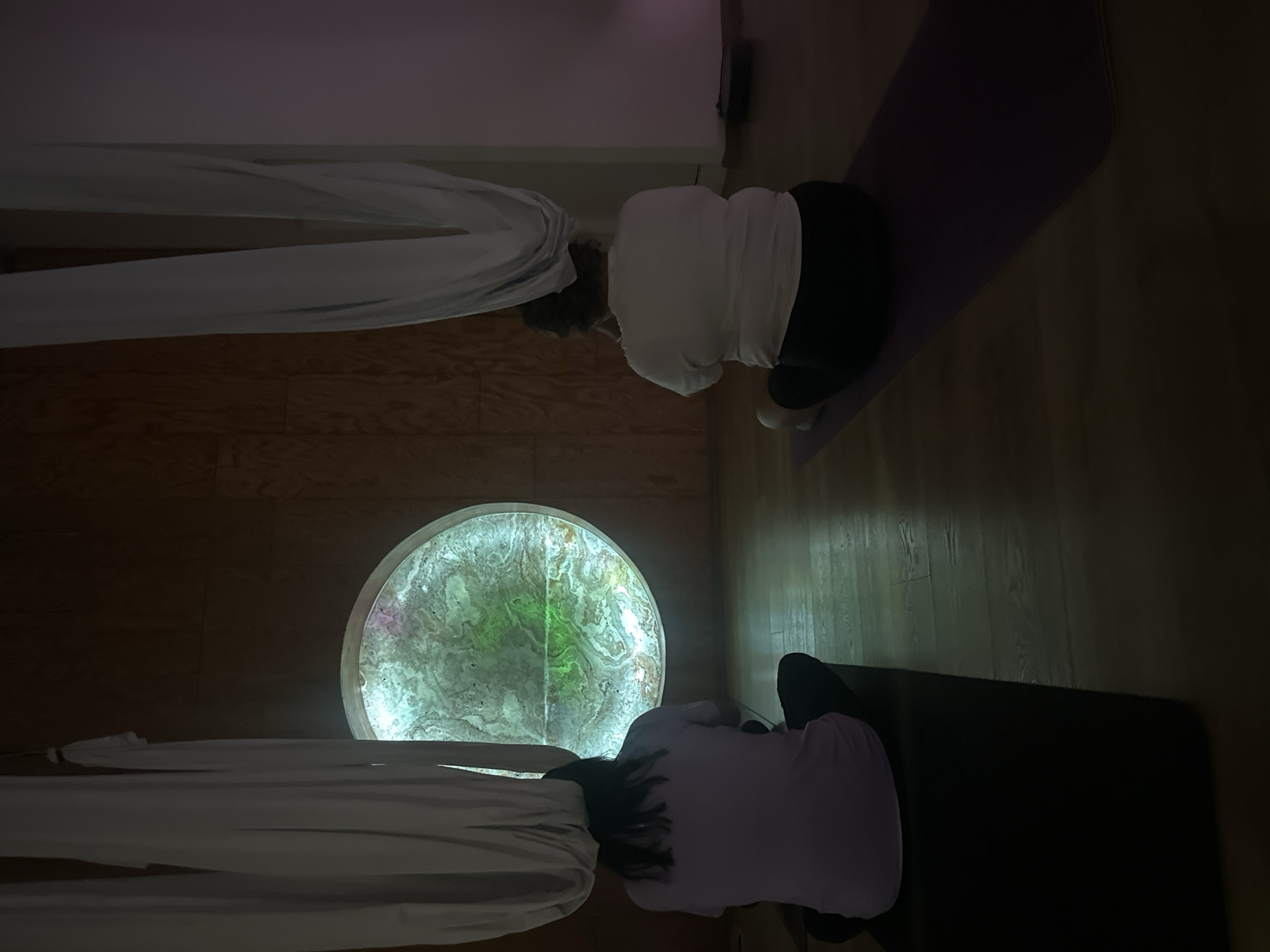 Two people are positioned on yoga mats in a dimly lit room with wood floors. One person wears a white shirt and black pants, while the other wears a lavender shirt and black pants. A round, illuminated panel is centered between them and the room appears to be in Puebla, Mexico. Two lengths of white fabric hang from the ceiling near the participants.