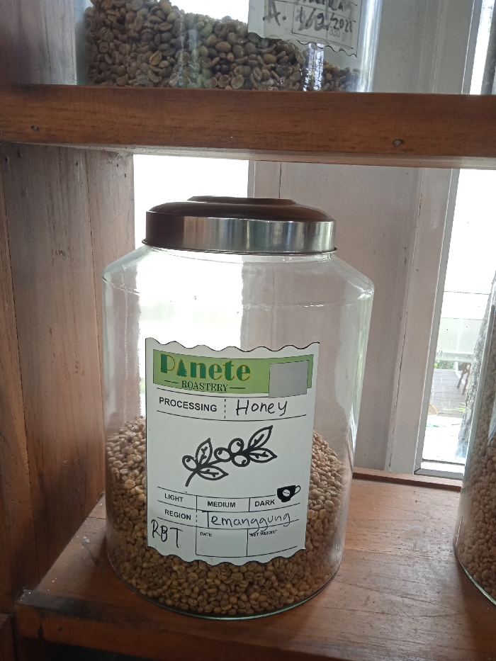 A glass jar filled with coffee beans sits on a wooden shelf in a coffee roastery called "Panete Roastery" in Plaosan, Indonesia. The jar has a silver lid and is filled with light to medium brown coffee beans. A white sticker on the front of the jar states "Processing: Honey, Region: Temanggung, RBT." The sticker is on a white background with green and black lettering. The jar is sitting on a wooden shelf, and there are more coffee beans visible in a jar above it and a jar to the right of it. The shelf is in front of a white-painted window. The window appears to be open to the outside, with the silhouette of a dog visible on a porch behind the window. The coordinates provided are located in the city of Plaosan, Indonesia. The scene appears to be a coffee roastery.