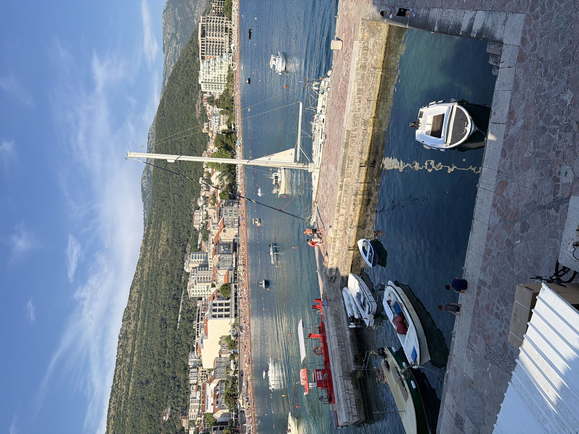 A wide-angle, vertical shot captures a sunny day at a marina in Petrovac centar, Montenegro. The foreground is dominated by a stone-paved dock with several small boats moored in the clear blue water. These include two white motorboats, one with a dark blue stripe, and a small, blue-hulled dinghy. Further out, a larger white sailboat with a raised mast is docked. Adjacent to the sailboat is a red rescue boat with "12-8 RD NAUTILUS" printed on its side. Two individuals, a man and a woman, are visible on the dock near the red boat, appearing to interact.
Beyond the marina, a crowded beach stretches along the coastline, lined with buildings that transition into a lush, tree-covered mountain range. Numerous small boats are scattered across the water between the beach and the marina. The sky above is a clear blue with sparse, wispy clouds. Two children are seated on the edge of the dock in the lower portion of the frame, while another individual stands on the far right of the dock, looking towards the water. The scene conveys a typical summer day at a coastal town.