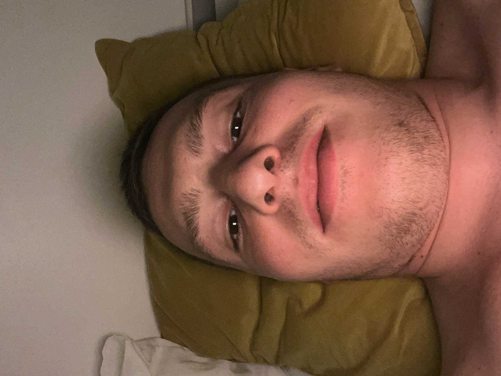 A close-up image shows a person lying down, with their head and upper chest filling the frame. The image is oriented with the person's head on the left side, as if looking upwards from a prone position. The subject has short, dark hair and visible stubble or light beard growth on their chin and upper lip. Their dark eyes are open and looking towards the camera, and a soft, contented smile plays on their lips.
The person's head rests on a soft, crumpled, greenish-yellow (olive-colored) pillow. The lighting is dim and warm, creating a relaxed atmosphere, possibly suggesting evening or nighttime. To the left of the person's head, a plain, light-colored wall or headboard is visible, while a white sheet or blanket can be seen beneath the pillow on the bottom right. The scene appears to be indoors, likely in a bedroom. There is no visible text in the image. The general location context is Vitry-sur-Seine, France.
