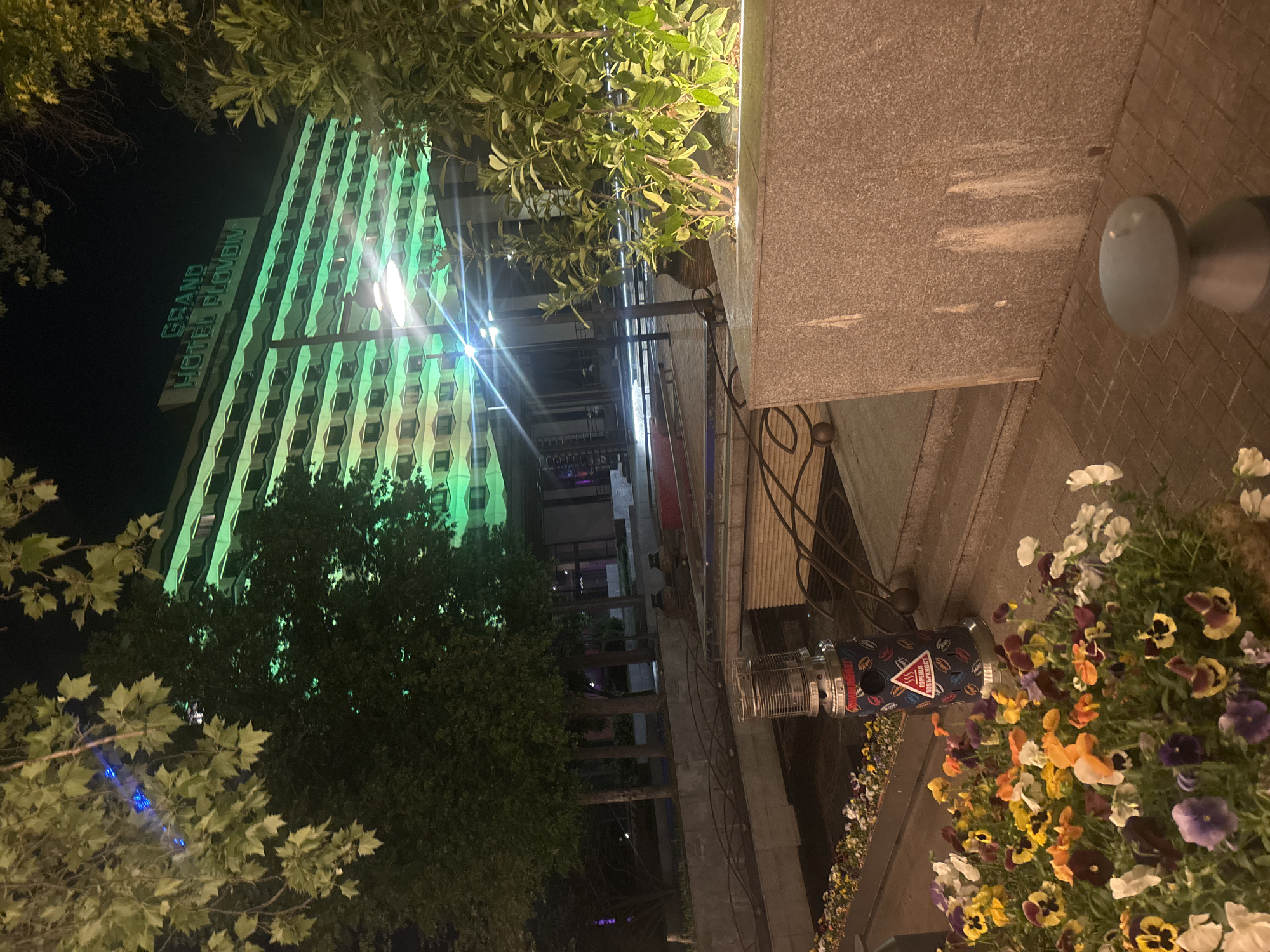 The image depicts an evening scene in Plovdiv, Bulgaria. The Grand Hotel Plovdiv is illuminated with green lights. In the foreground is a sidewalk with flower arrangements and a patio heater. People are not visible in the image. The scene seems to be taken from a pedestrian area.