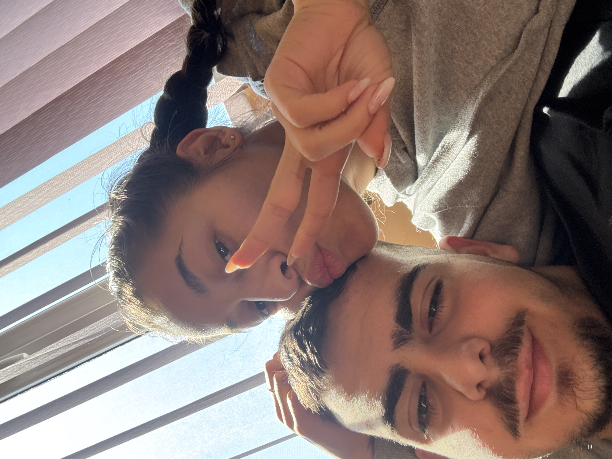 Two people are in the image. A woman with dark hair pulled back in a braid makes a peace sign with her fingers. Her face is tilted to the side as she is laying on top of a man with dark hair, a mustache, and a beard. They are indoors. The background includes window blinds. The location is not recognizable as being in Plovdiv, Bulgaria.