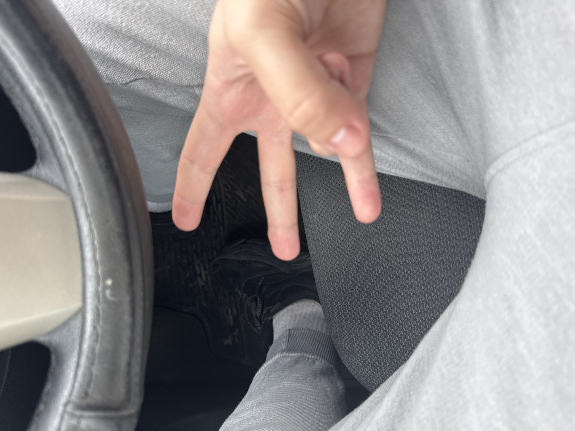 A close-up photograph shows a hand displaying three extended fingers, positioned above the lower half of a person seated in a vehicle. A steering wheel is visible on the left. The person is wearing grey sweatpants and a sock, with a black shoe on the floor. The seat is dark grey. The location is not discernible from the image, so the image does not provide enough information to determine whether it was taken in Plovdiv, Bulgaria.