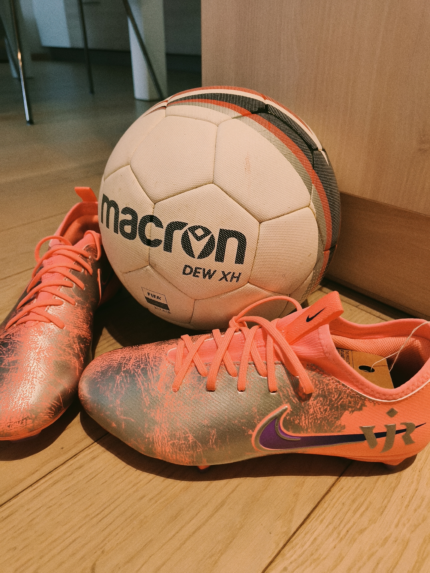 A close-up shot shows a soccer ball and a pair of soccer cleats resting on a light-colored wooden surface. The soccer ball is white with the brand name "Macron" in black bold letters and "DEW XH" underneath. The ball also has black, gray, and red striped accents near its seams. The soccer cleats are primarily coral or salmon-colored with silver textured designs on the front part of the shoe. They have coral-colored laces and a purple Nike swoosh on the sides. The shoes also feature the letters "VJ" in silver near the heel. A price tag is visible inside the right cleat. The scene is indoors, likely in a home or retail setting. It is not possible to ascertain from the given image that the photograph was taken in Sofia, Bulgaria.