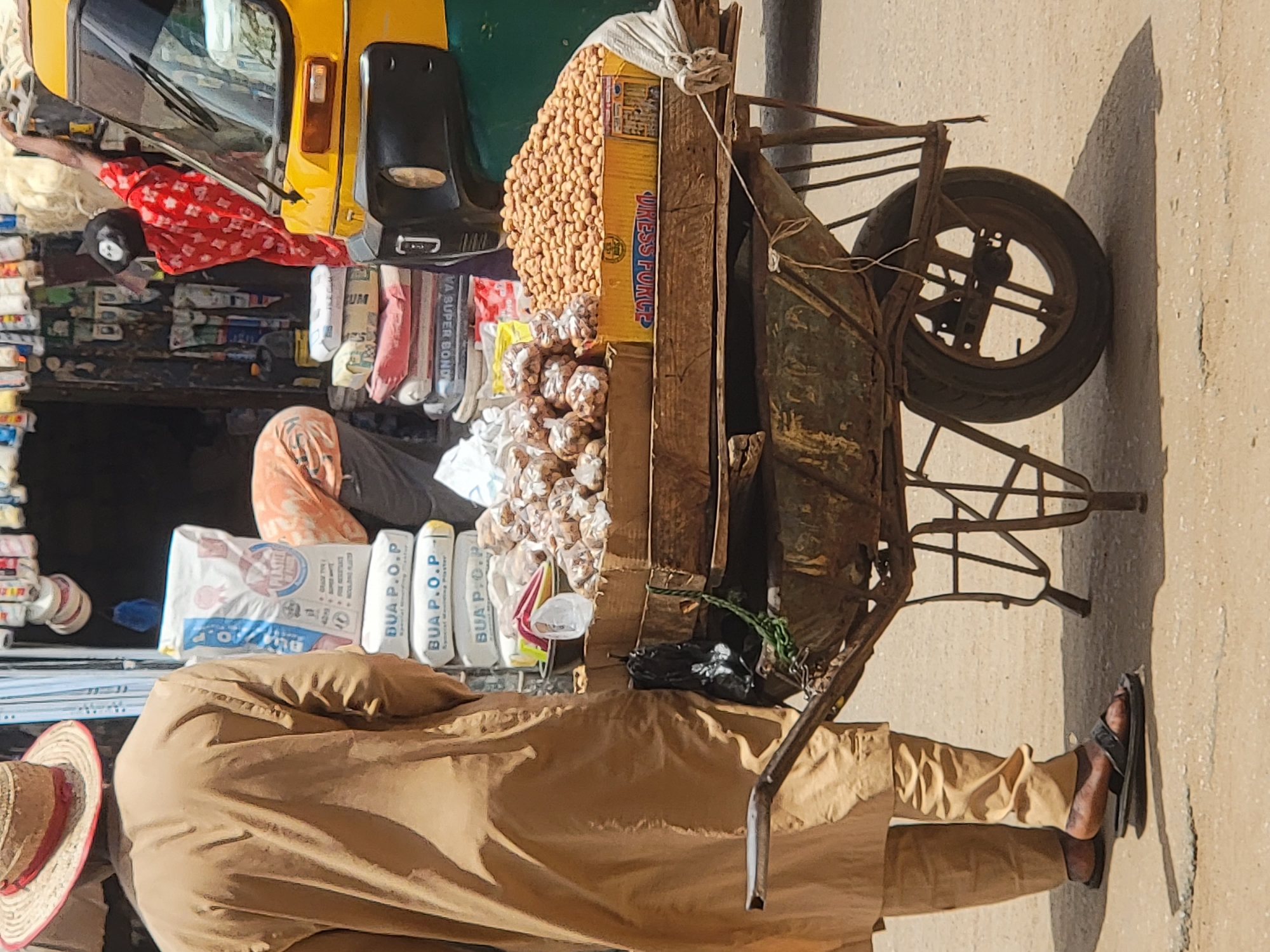 An outdoor scene in Maiburiji, Nigeria, depicts a street vendor and their pushcart laden with goods on a sunny day. The image is taken from a slightly elevated perspective, looking down at the street level.
In the foreground, a person, likely the vendor, is visible from the waist down, wearing light brown or khaki-colored loose clothing and black sandals. A straw hat with a red-striped brim rests near them on the left side. They stand next to a rustic, dark-colored metal pushcart with one visible spoked wheel and a rubber tire.
The pushcart is filled with merchandise. On top, a large quantity of light brown, oval-shaped items, likely groundnuts (peanuts), are piled onto a yellow cardboard box. Below these, numerous smaller bundles, possibly garlic or other produce, are individually wrapped in clear plastic. A black plastic bag and some green leafy vegetables are also visible on the cart. The yellow cardboard box has the partially visible text "LIKESS FORCE" and a logo.
In the background, behind the vendor and cart, a bright yellow auto-rickshaw (keke Napep) is partially visible, parked or driving on the street. Several individuals are in the mid-ground: one person in a red and white patterned garment, another with an orange head covering, and another wearing a black cap. Along what appears to be a building or stall wall, various white and grey sacks are stacked. Visible text on these sacks includes "BUA P.O.P." (Special Gypsum Plaster) and "MA SUPER BOND". The ground is paved, possibly asphalt or concrete, and the strong shadows indicate bright, direct sunlight. The scene captures a bustling street or market environment.