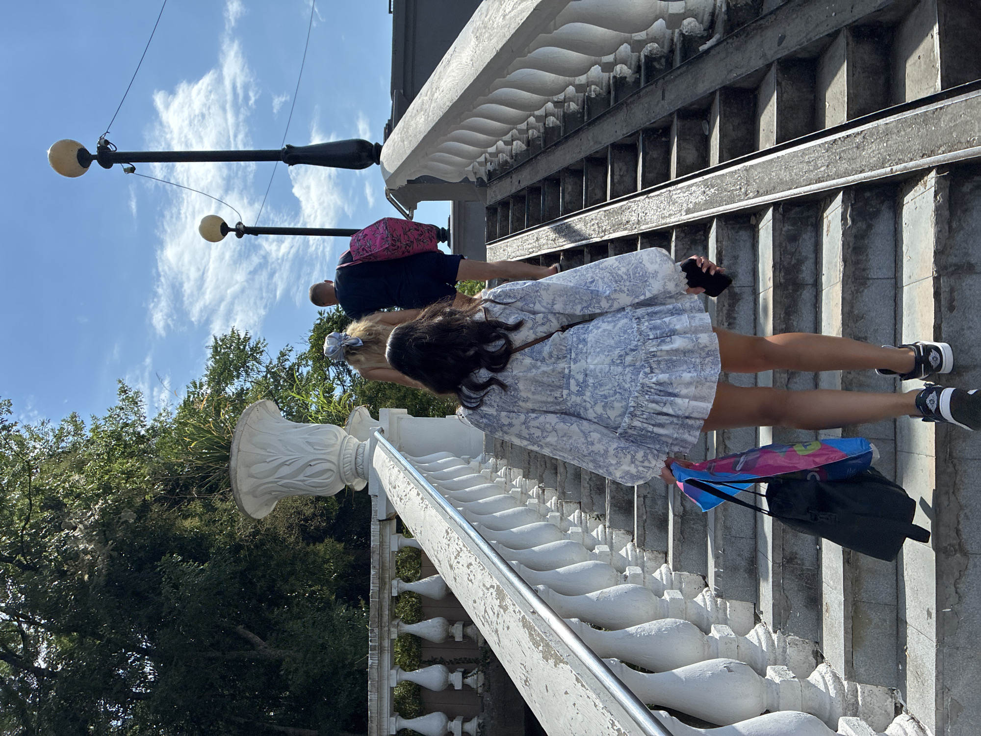 Three individuals are ascending a wide outdoor stone staircase with white balustrades. The person in the foreground, a woman with dark hair wearing a blue and white patterned dress and black sneakers, carries a black duffel bag and two brightly colored shopping bags in her right hand, and a dark-colored smartphone in her left hand. To her left, a man in a dark t-shirt and navy shorts carries a large pink bag over his right shoulder. Further ahead, a woman with blonde hair, wearing a light-colored scarf as a headband, walks ahead of the man. The staircase is bordered by lush green trees on the left and features a metal ramp alongside the right side of the stairs. The sky above is blue with scattered white clouds, and two tall lampposts with globe-shaped lights are visible against the sky. This scene is likely located in Sochi, Russia, given the architectural style and vegetation typical of the region.