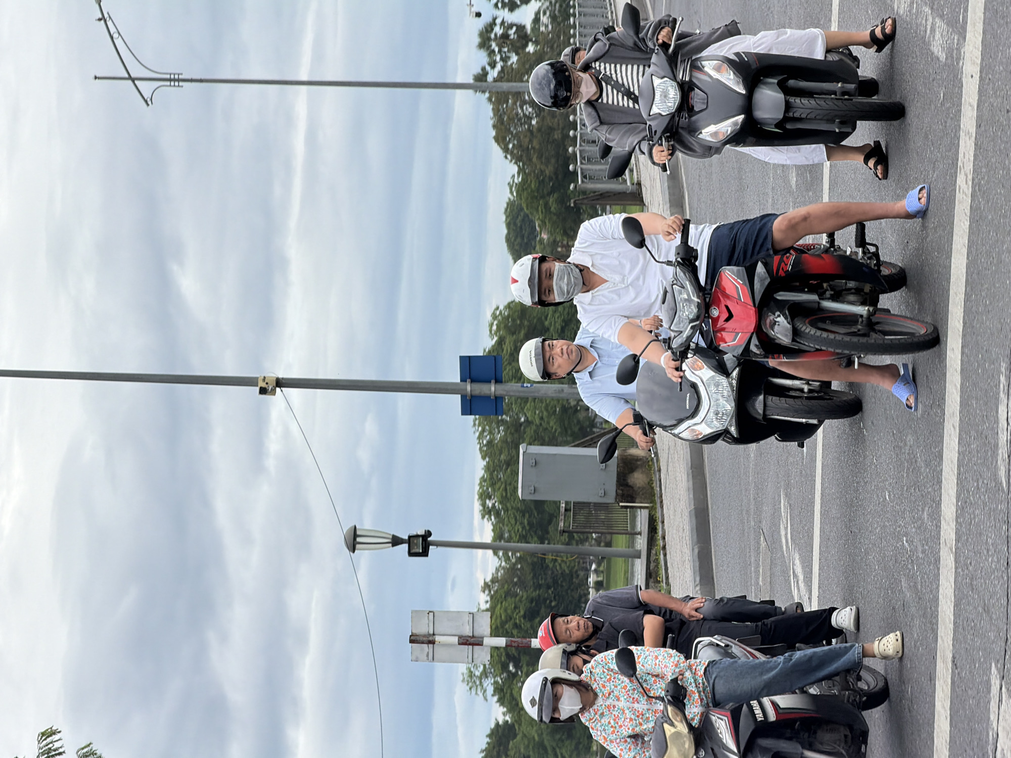 In the city of Thuận Hóa Ward, Vietnam, under a cloudy sky, a group of individuals on motorcycles waits at an intersection. The foreground features three motorcycles, with riders wearing helmets and face masks. On the left, a woman in a floral shirt and jeans is on a Yamaha scooter, accompanied by a man behind her wearing a dark shirt and a red helmet. To her right, a man in a light blue shirt and a white helmet is on a motorcycle, also wearing a face mask. Further to the right, a man in a white polo shirt and dark shorts, wearing a white helmet and face mask, rides a red and black motorcycle. The fourth motorcycle, on the far right, is ridden by a person in a grey striped jacket and white pants, wearing a black helmet and a pink face mask. The background shows trees, a street lamp, a traffic light pole, and a barrier. The road surface is asphalt.