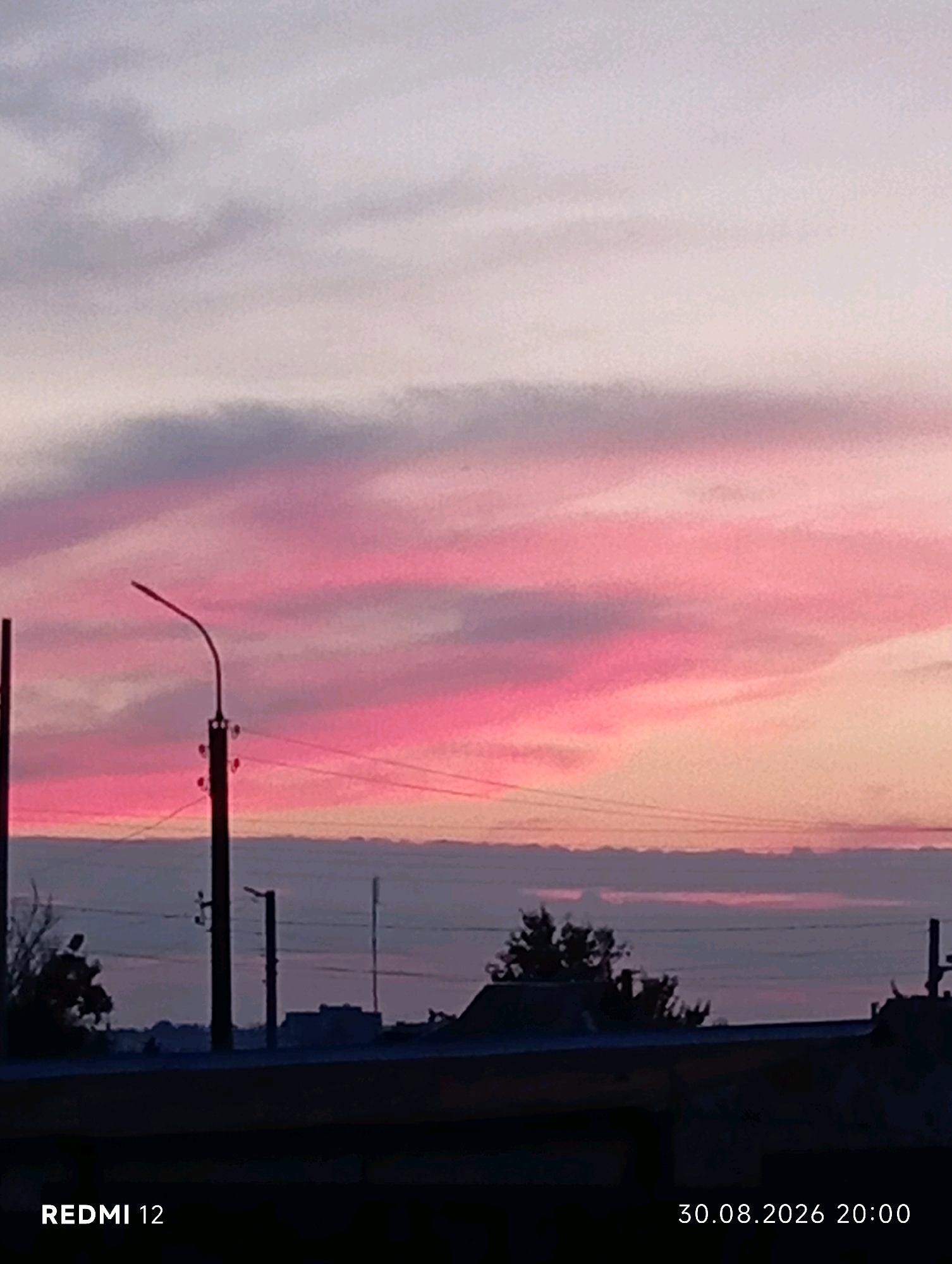 The image captures a dusk sky with vibrant pink and gray clouds, hinting at a sunset. In the foreground, silhouetted against the colorful sky, are the outlines of buildings and trees. A street lamp with its curved arm is prominent on the left side of the frame, along with power lines stretching across the scene. The bottom left corner of the image displays "REDMI 12," and the bottom right corner shows "30.08.2026 20:00," indicating the device used and the date and time the photograph was taken. The general environment suggests an urban or suburban setting, likely in Smila, Ukraine, based on the provided context.
