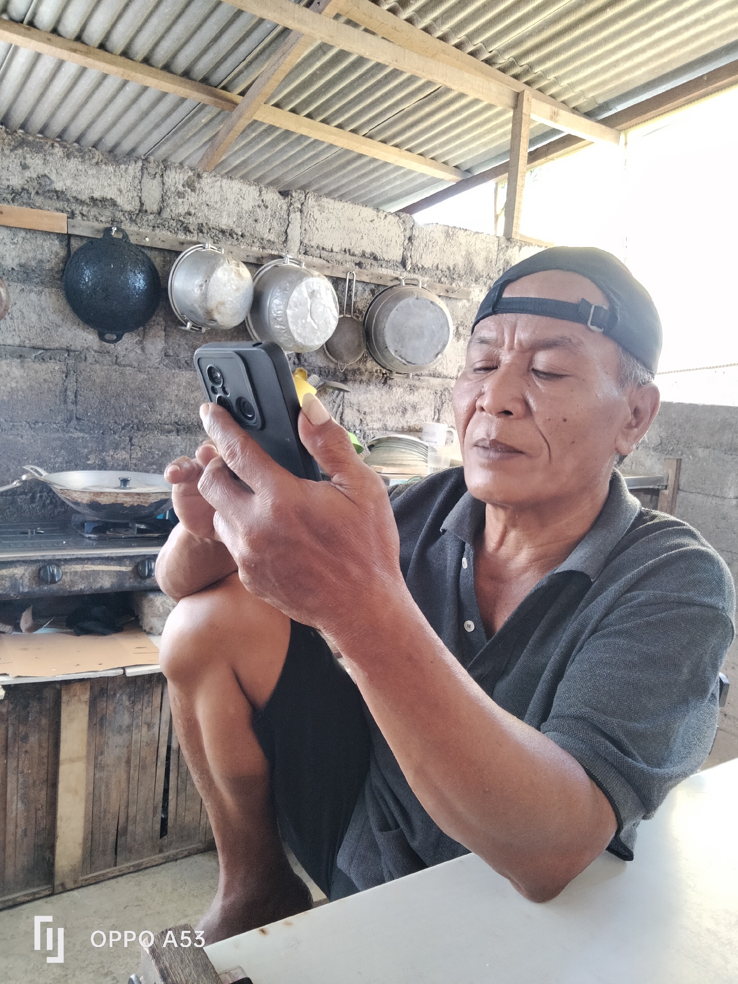 A man wearing a gray polo shirt and a black baseball cap sits on the floor, looking at his phone. He is in what appears to be an outdoor kitchen area with a corrugated metal roof supported by wooden beams. The background includes a stone wall with several pots and pans hanging from it, and a stovetop with a pan on it. The floor is made of concrete. A white table is in the foreground. The image appears to be taken in Denpasar, Indonesia.