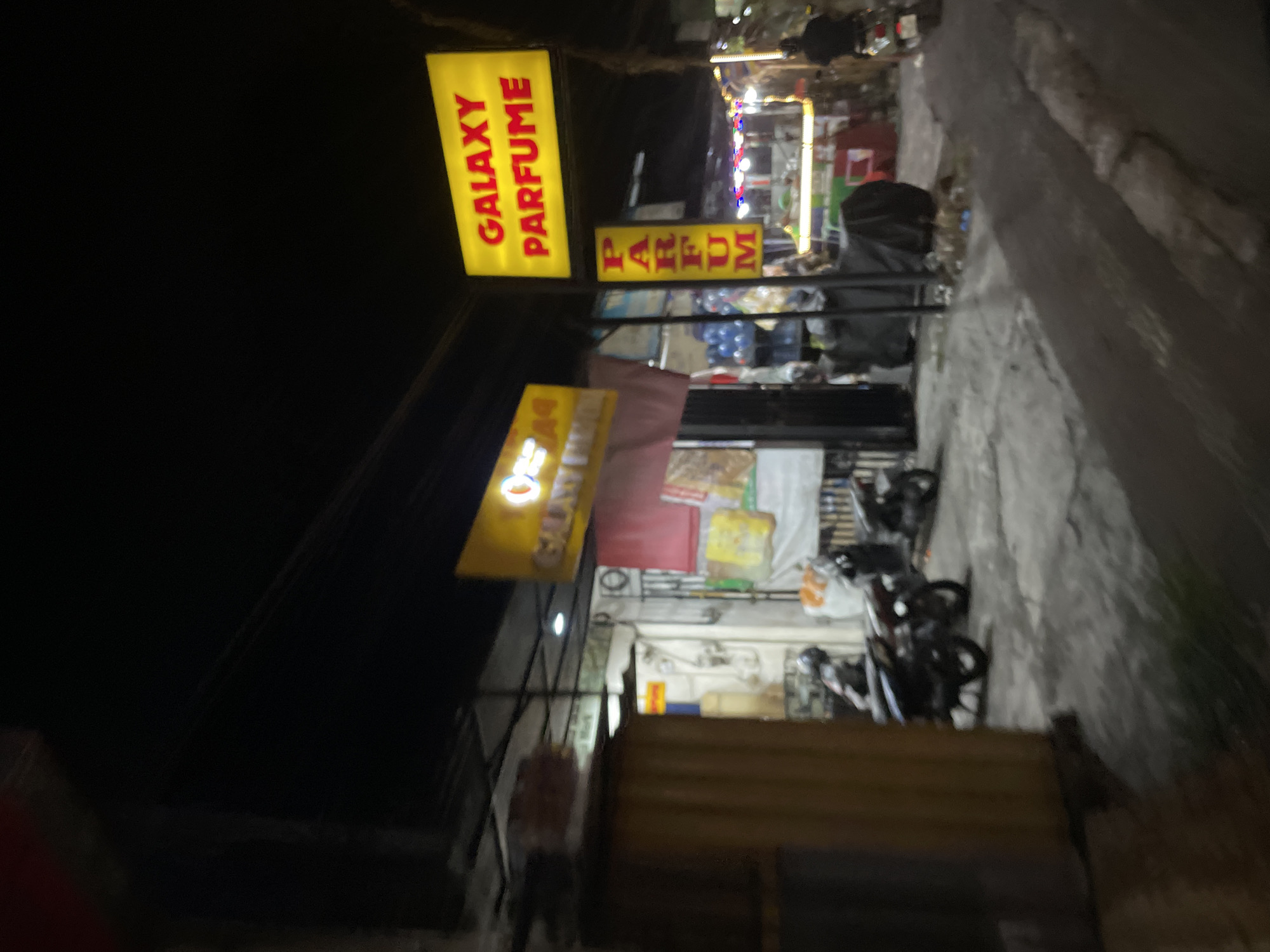 The image shows a street scene at night in Denpasar, Indonesia. A bright yellow sign with red lettering, which reads "GALAXY PARFUME," is prominently displayed on the left side of the frame. Below it, another yellow sign with red lettering reads "PARFUM." In the background, a store or market is visible with lights illuminating the interior. Several motorcycles are parked in front of the establishment. The overall scene suggests a commercial area with shops operating late into the night. The lighting is dim, indicating it is nighttime, and the weather appears to be clear. The image is slightly blurry, which could be due to low light conditions or movement during the capture.