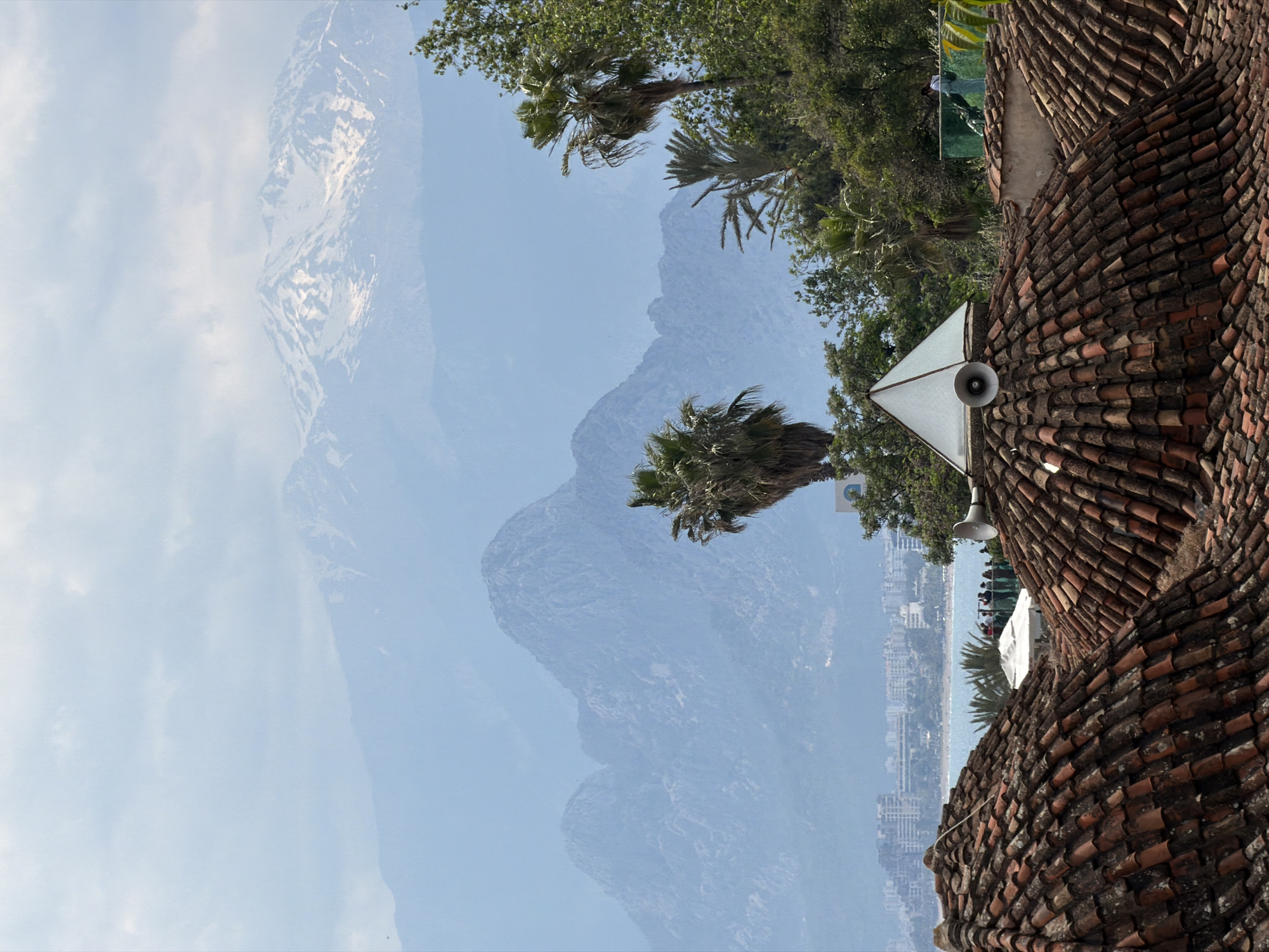 The image is taken outdoors in Antalya, Turkey. The frame is split into two primary sections: the left side features an obscured mountain range against a bright sky, and the right side showcases a rooftop covered in reddish-brown tiles. Palm trees are sporadically visible. There is a white, pyramid-shaped structure on the roof, equipped with a loudspeaker. In the far distance, a cityscape lines the coast, appearing between the rooftop and the trees. A group of individuals can be seen at a viewing point.
