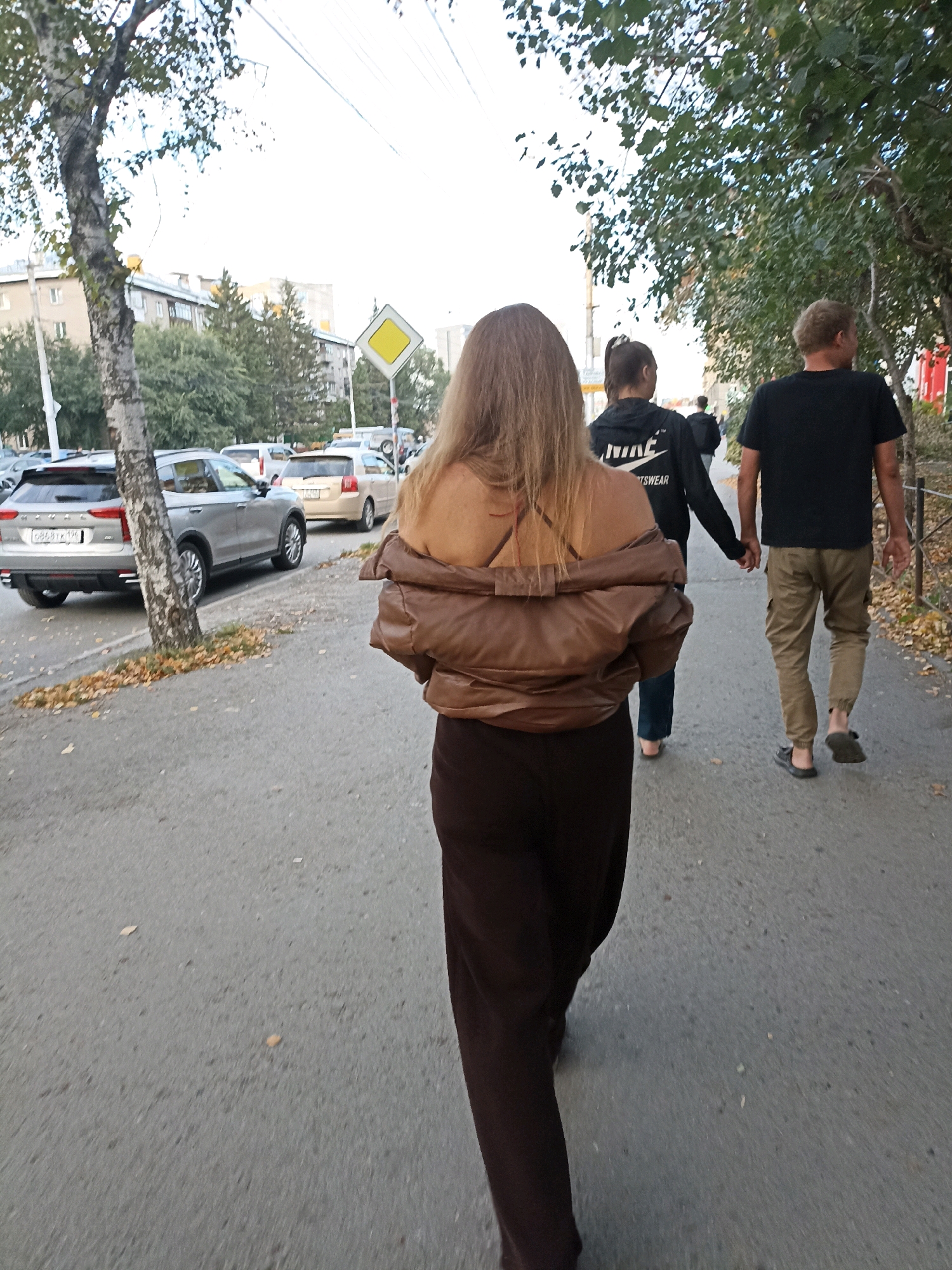 Three people walk along a paved sidewalk. In the foreground, a person with long blonde hair wears a brown puffer jacket over their shoulders and dark brown pants. Behind them, a second person, seen from the back, wears a black hoodie with "NIKE SPORTSWEAR" printed on it, dark pants, and is barefoot. They hold hands with a third person, a man with short blonde hair, wearing a black t-shirt and tan cargo pants. He is also barefoot and wears dark flip-flops. The sidewalk is next to a street with several parked cars, including a silver SUV. Trees with green and yellowing leaves line the sidewalk and street. In the background, a yellow diamond-shaped traffic sign is visible. The sky is bright and overcast.