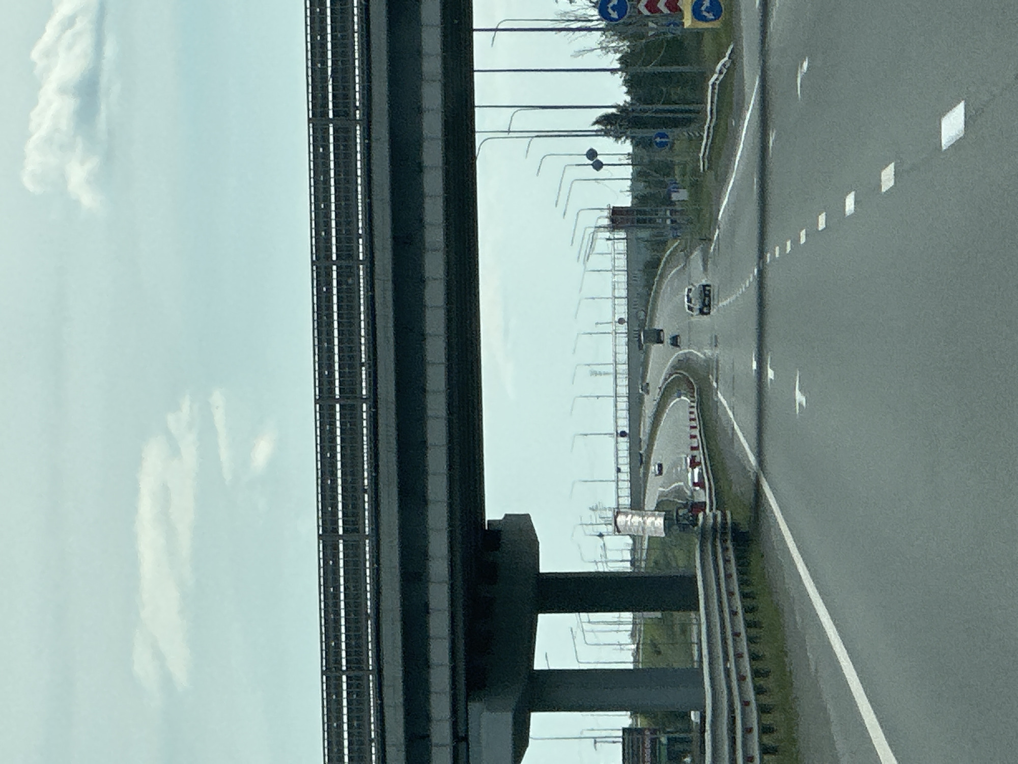 The image shows a view from a car traveling on a multi-lane highway beneath an overpass. The highway is paved and features white lane markings. Several cars are visible on the road, some in the foreground and others further in the distance. The overpass is constructed of concrete and has a metal railing on top. The sky is overcast with scattered clouds. Streetlights line the sides of the highway. Traffic signs are also visible, including directional signs and regulatory signs. The environment appears to be an urban or suburban area, with some greenery visible along the sides of the road.