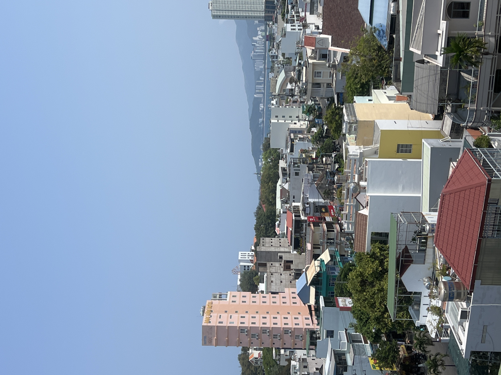 The image depicts a dense urban area in Nha Trang, Vietnam, characterized by tightly packed buildings of varying heights and colors. Predominantly visible are residential structures, likely houses and apartments, with a mix of architectural styles. Many rooftops are accessible, featuring satellite dishes, water tanks, and makeshift enclosures. Greenery is interspersed amongst the buildings, with trees and other vegetation dotting the landscape. In the distance, a hazy mountain range forms a backdrop to the city, with a taller skyscraper standing out from the horizon. The sky is clear and blue, indicative of a bright day. A pink hotel building can be seen in the foreground, with signage visible. The overall scene suggests a bustling, densely populated area.