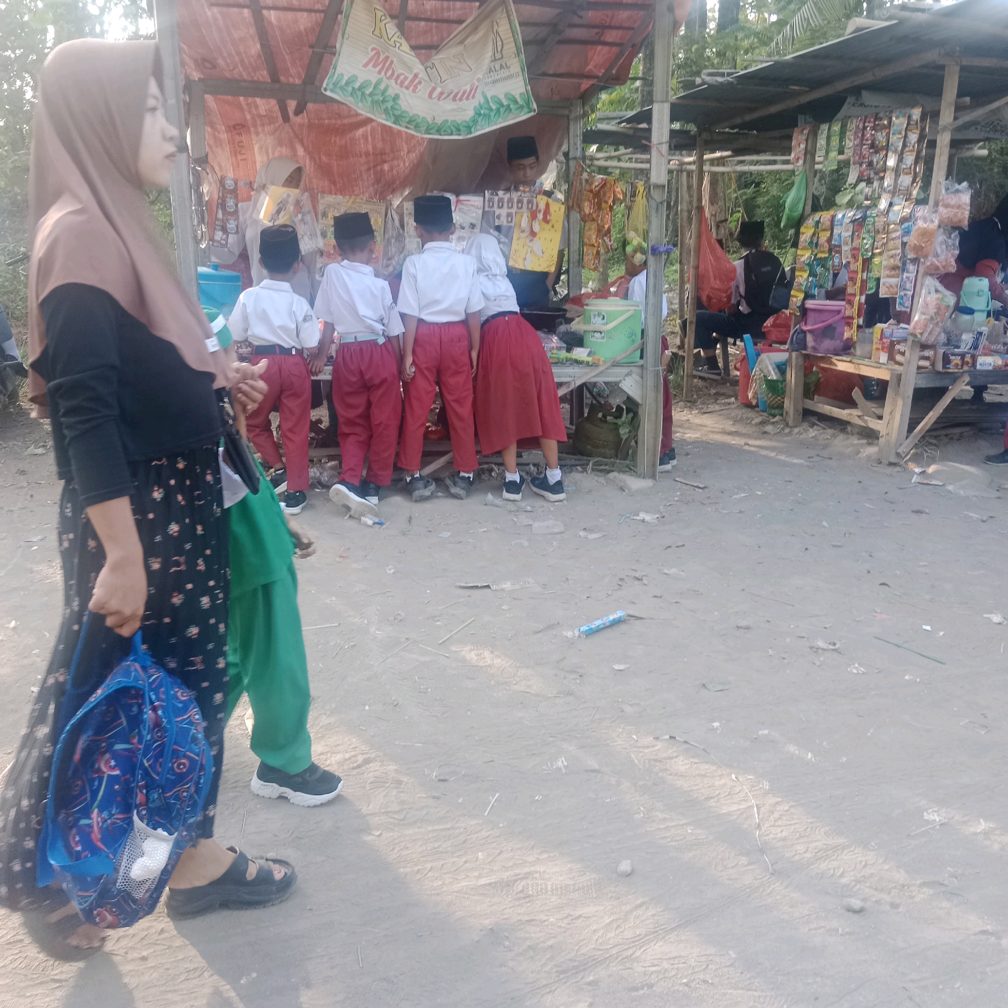 The image depicts an outdoor scene at what appears to be a small roadside market or stall in Tegowanu Kulon, Indonesia. In the foreground, a woman wearing a brown hijab and a patterned black dress is walking, carrying a blue backpack. Her left hand is visible, and she is wearing black sandals. To her right, several children in school uniforms, consisting of white shirts and red pants or skirts, are gathered around a stall. A young boy in a black cap is standing behind the stall, appearing to be the vendor, interacting with the children. The stall itself is constructed of wood and metal, with a corrugated metal roof covered by a tarp. It is stocked with various packaged snacks and goods. Behind the stall, trees and foliage are visible, suggesting a rural or semi-rural setting. The ground is dirt and gravel, with some scattered debris. Sunlight casts shadows across the scene, indicating it is daytime.