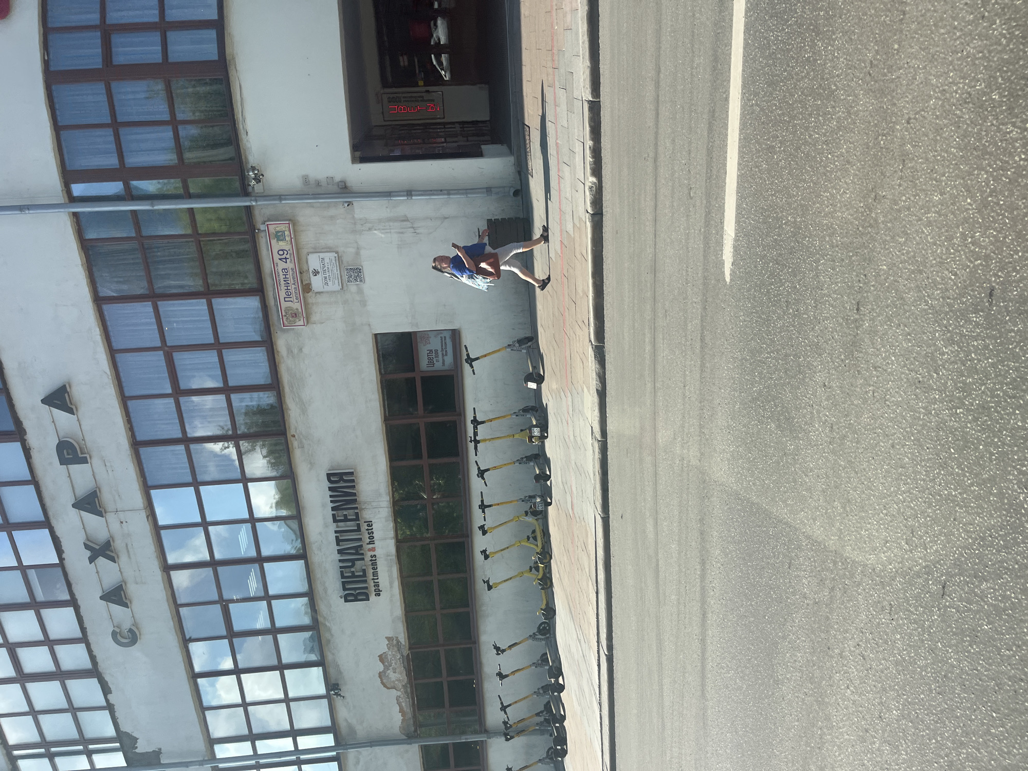 In Yekaterinburg, Russia, on a sunny day, a woman walks along a sidewalk in front of a building with large windows and Cyrillic lettering that reads "SAHAPA" and "VPЕЧАТЛЕНИЯ apartments & hostel." A street sign indicates "Lenina Avenue 49." A row of yellow electric scooters is parked on the sidewalk. The woman, wearing a blue top and light-colored pants, carries a bag over her shoulder and looks at her phone. To the right, an entrance to a building is visible with a sign that says "ЦВЕТЫ," meaning "flowers." The street in the foreground is paved and has a white line marking.