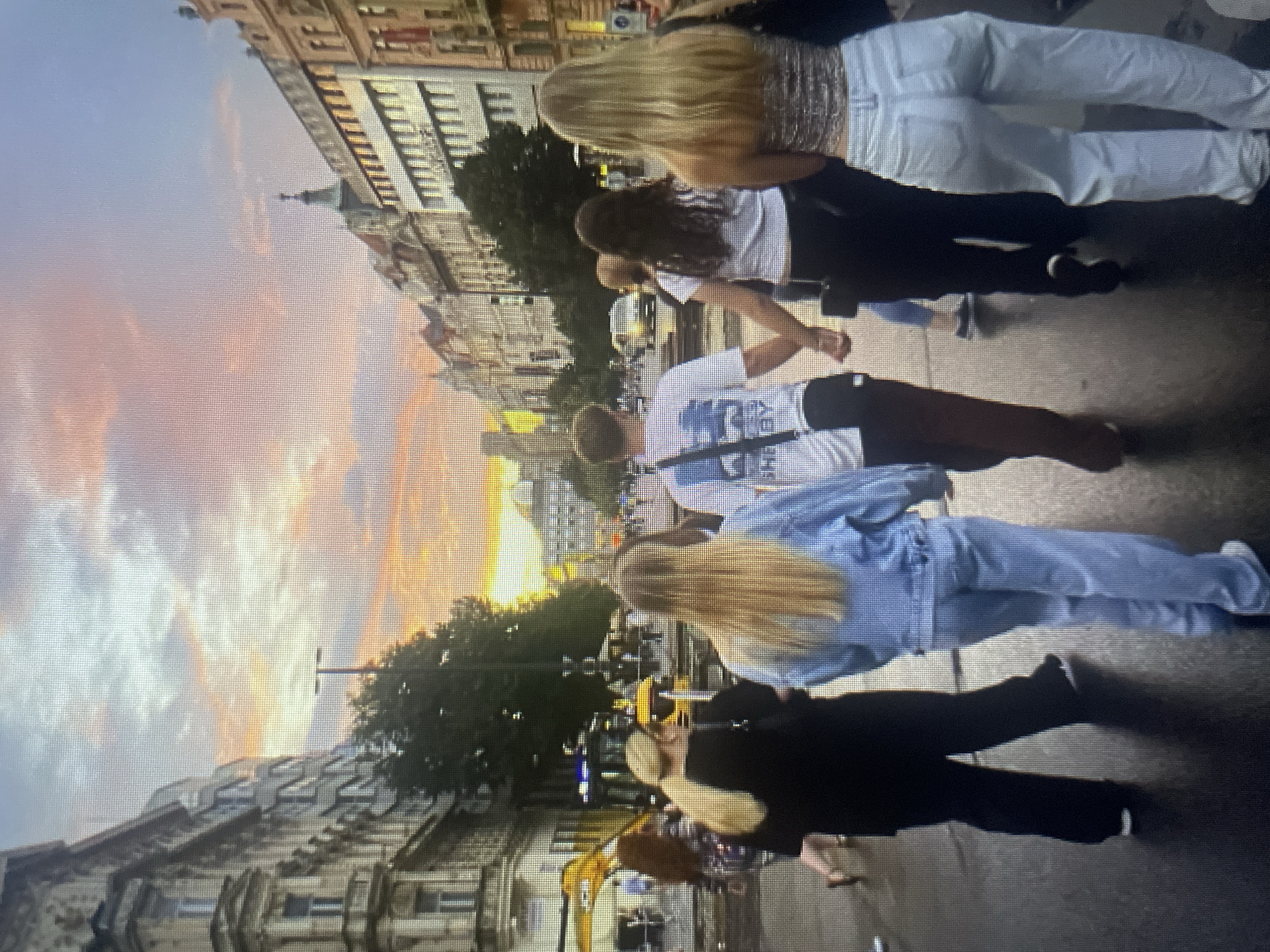 In an urban environment, under a dramatic sunset sky with hues of orange, pink, and blue, a group of five individuals, four females and one male, are captured from behind, walking across a street. The street appears to be a thoroughfare with vehicular traffic and is lined with multi-story buildings.
The individuals are varied in their attire. Two women in the foreground are wearing light-colored denim. One is in a denim jacket and matching light blue jeans, while the other is in a sparkly patterned top and light blue jeans. A third woman, to the left, wears a black top and black pants. A fourth woman, also in the foreground but partially obscured, has dark hair and is wearing a white crop top and dark pants, holding hands with the male. The male is wearing a white t-shirt with a graphic print and dark shorts, and has a dark bag slung across his chest. A fifth individual with dark hair and a patterned top is walking further in the background to the left.
The environment features architectural elements of city buildings, trees, streetlights, and what appears to be a tram or streetcar track on the road. The lighting suggests it is late afternoon or early evening.
While the image depicts a European city street scene at sunset, there is no visual information within the image to indicate it is specifically located in Kołobrzeg, Poland.