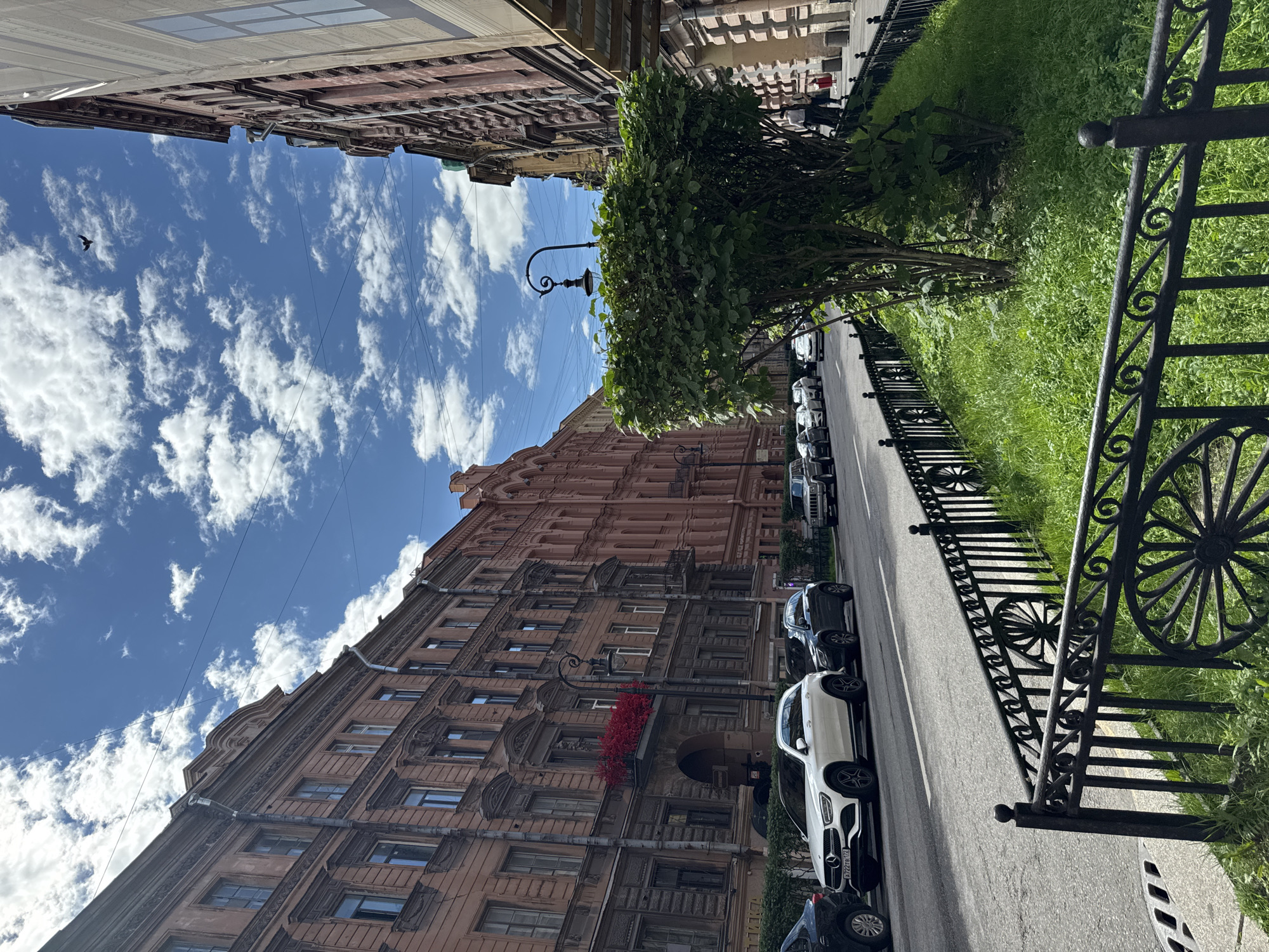 The image depicts a street scene in Saint Petersburg, Russia. The perspective is from ground level, looking upwards at buildings lining a street. The buildings are tall, multi-story structures with ornate architecture, characteristic of historical European cities. One building is a pale pink color, while others appear to be a reddish-brown or grey stone. The sky is a vibrant blue with scattered white clouds, suggesting a pleasant daytime, likely late morning or afternoon.
Along the street, several cars are parked. A white Mercedes-Benz is prominently featured, along with other dark-colored vehicles. A black metal fence with decorative scrollwork separates the sidewalk from a strip of lush green grass and a tree. The tree is full and leafy, adding a touch of nature to the urban environment. A traditional street lamp with a curved arm stands near the tree.
A small detail of bright red flowers is visible on a balcony of one of the buildings, providing a pop of color against the muted tones of the architecture. A bird is visible in the sky, a small dark silhouette against the clouds. The presence of overhead wires suggests an established urban setting. The overall impression is of a well-maintained, historic city street on a clear day. No specific activity or event is taking place, rather it is a snapshot of everyday life. There is some visible text on the license plate of the white Mercedes-Benz, but it is not legible.
