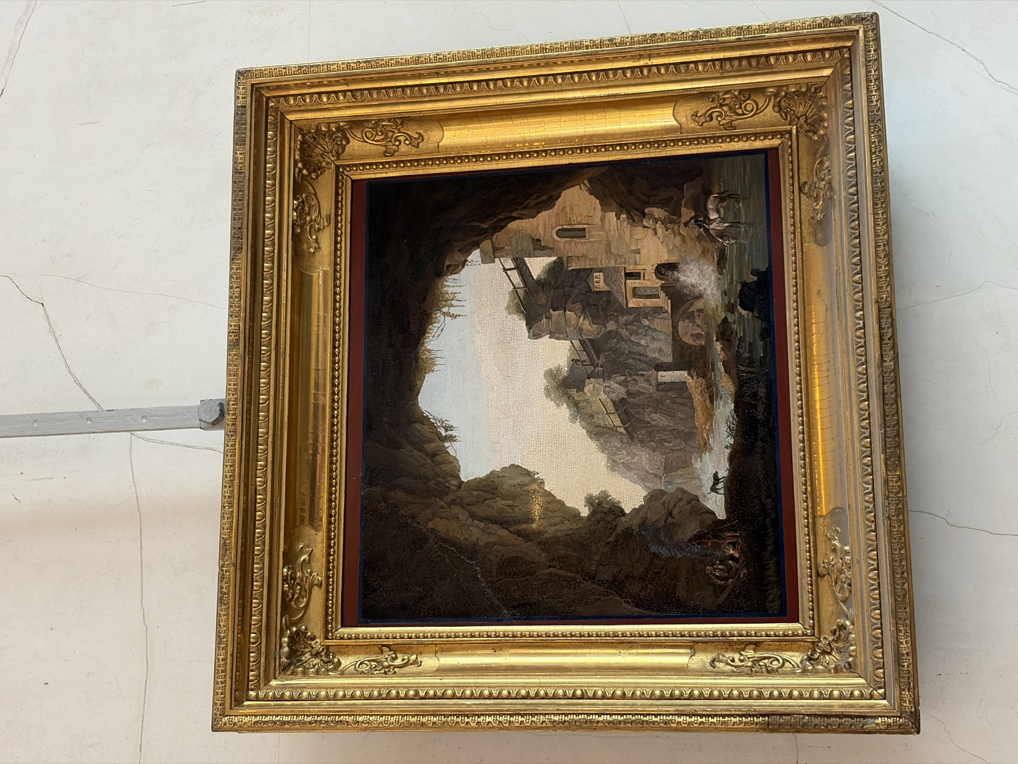 A framed painting of a mountainous landscape with buildings is hung on a white wall. The painting is in a heavy, ornate gold frame. The landscape features rugged cliffs, a waterfall, and stone buildings built into the cliffs. There are figures in the painting, including people near the waterfall and on the cliffs. The painting appears to be displayed in a gallery or museum setting. The location is Unknown, Unknown.