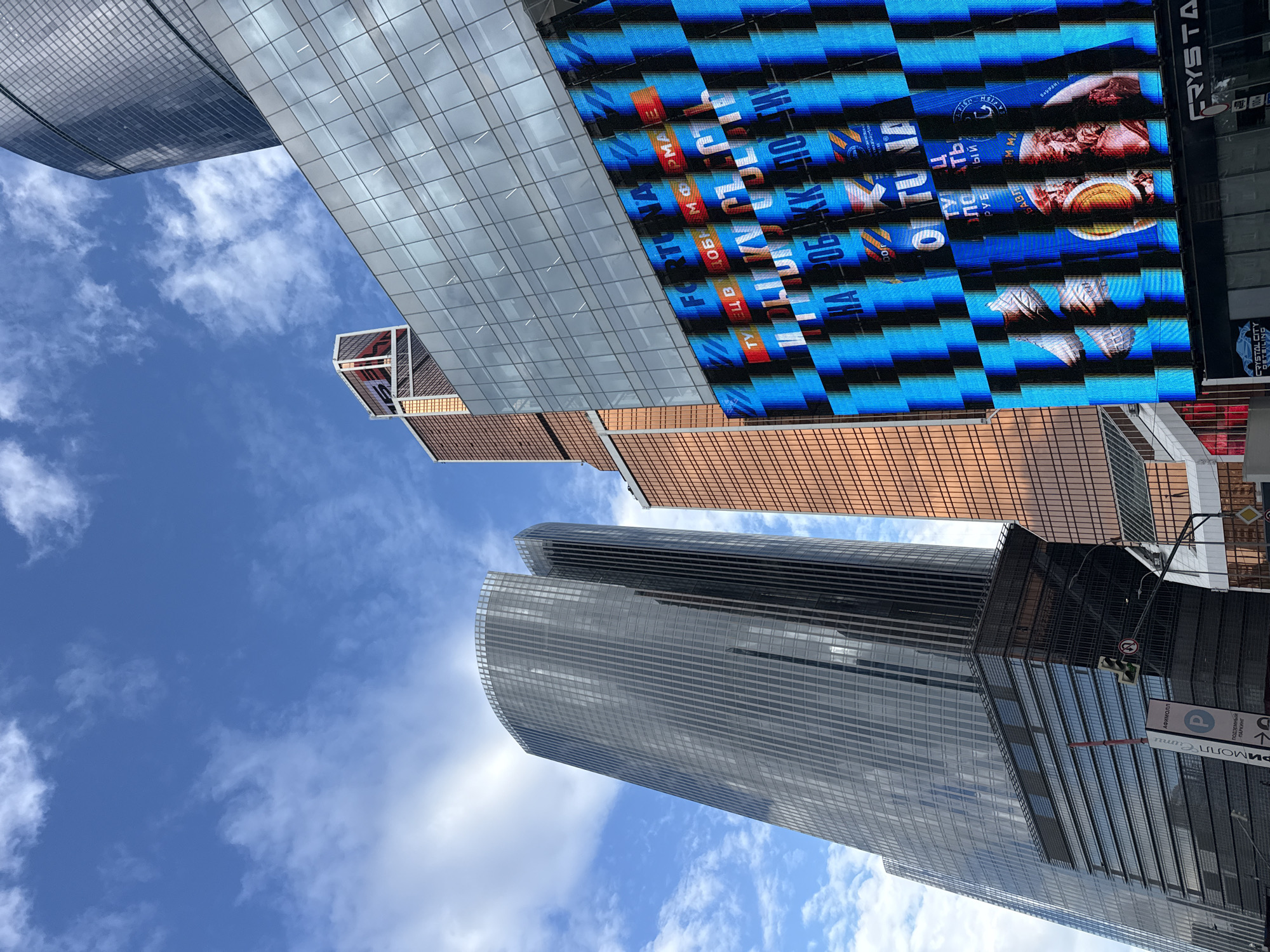 The image is an upward-angled shot of skyscrapers in Khimki, Russia, under a blue sky with scattered clouds. Three prominent skyscrapers dominate the frame. One is a tall, gray, cylindrical building with vertical lines. Another building has a reflective, copper-colored facade with a grid pattern. A large LED advertising display shows various images and text, including the words "FORTUNA," "ТУЕЦ," and what appears to be food items like soup. Other objects include street signs and traffic signals, indicating an urban environment. The buildings have glass exteriors.