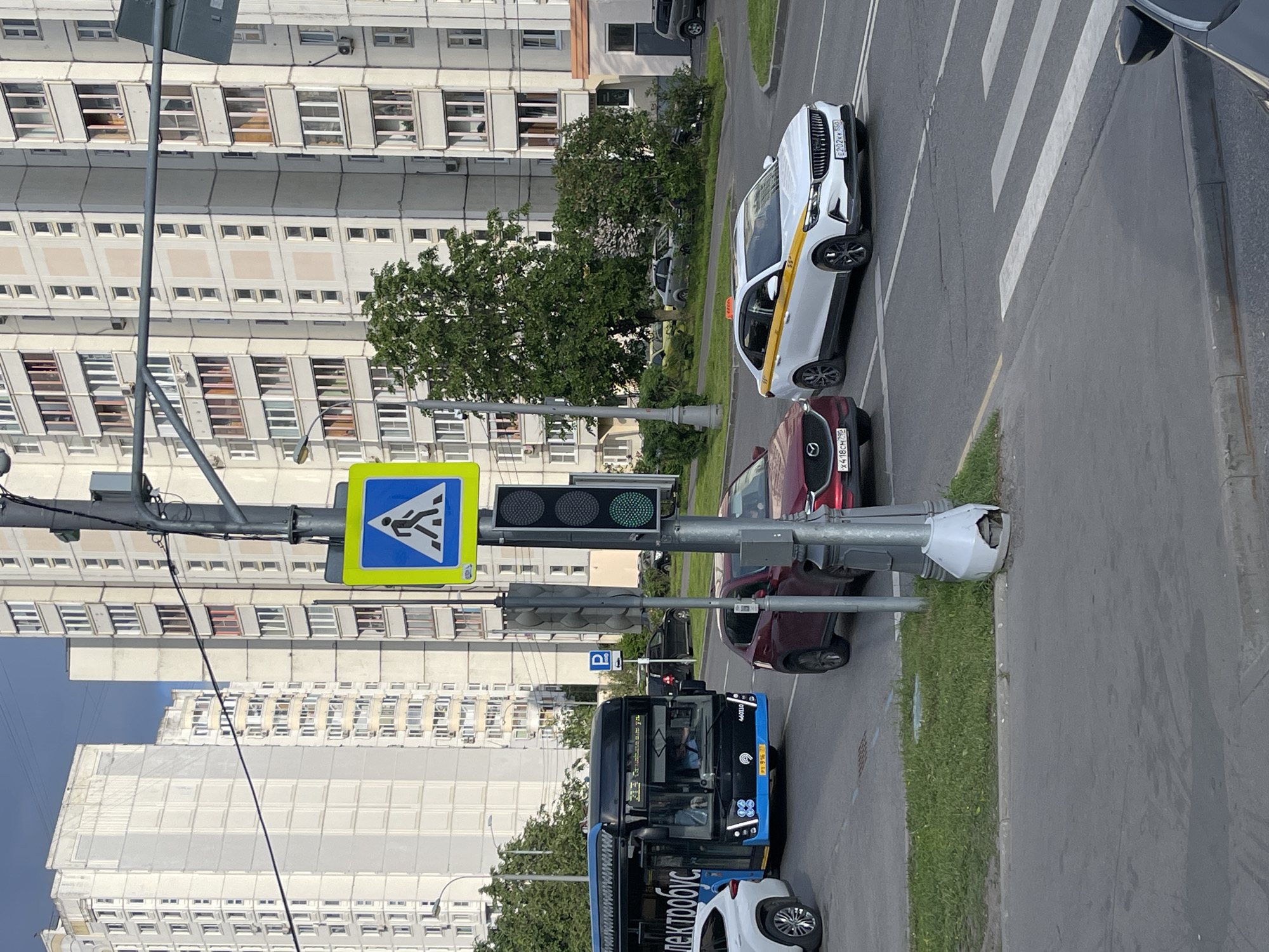 The image captures a street scene in Moscow, Russia, during the daytime under clear weather conditions. The perspective is from an elevated position, looking down onto a road with multiple lanes of traffic.
In the foreground and midground, several vehicles are visible. A white taxi, likely a Volvo, with a distinctive yellow stripe along its side, is prominent on the right, displaying the license plate E207XK 799. Behind it is a red Mazda car with the license plate X418CM 799. To the left, a blue electric bus, marked "электробус" (electrobus) and bearing the branding "MOCЭЛЕКТРОТРАНС" (Mosgortrans), is in view, with a driver visible inside. The bus also shows the number 460130. These vehicles appear to be in traffic, possibly at a stoplight or moving slowly.
A pedestrian crossing sign, a blue square with a white walking figure, is affixed to a traffic pole in the center-left. Below it, a traffic light displays a green signal, indicating that traffic can proceed. Further down the street, zebra stripes for a pedestrian crosswalk are visible on the asphalt. A small blue parking sign (P) is visible behind the bus.
In the background, a series of large, multi-story apartment buildings with numerous windows line the street. These buildings are characteristic of Moscow's residential architecture. Green trees and grassy areas separate the road from the buildings. The sky is bright and blue.