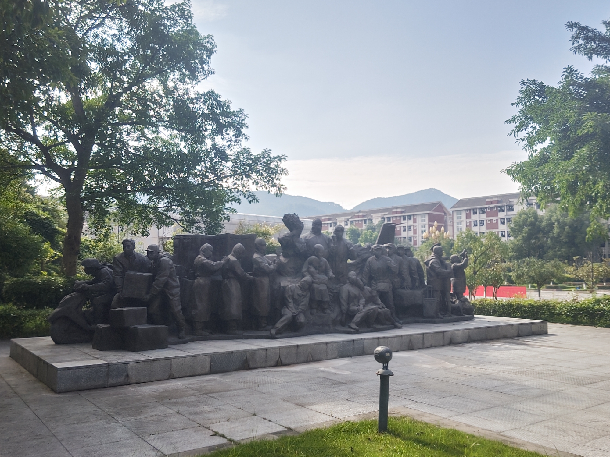 The image depicts a large, dark-colored bronze sculpture of a group of people engaged in various activities, including carrying boxes, riding a scooter, and conversing. The sculpture is situated on a raised, multi-tiered stone pedestal, which is surrounded by paved ground. To the left of the sculpture, a large tree with dense green foliage is prominent. In the background, several multi-story buildings with red roofs and white facades are visible, interspersed with more greenery and trees. Distant hills are also visible under a bright, clear sky. A small, dark lamppost stands on the paved area in the foreground, to the right of the sculpture. The overall scene suggests an outdoor public space, possibly a park or a plaza, within an urban environment. The location of this sculpture has been identified as being within the Chongqing High-tech Zone, China.