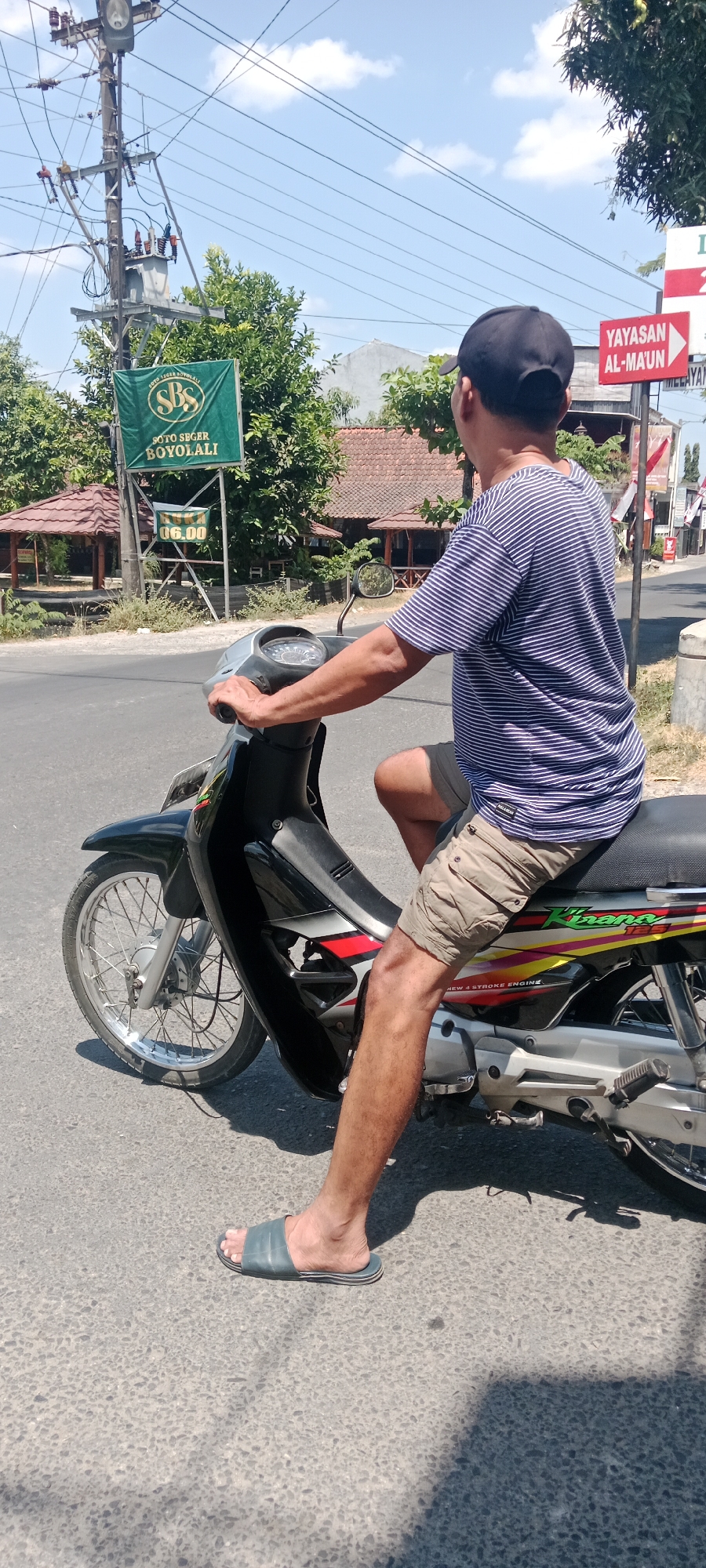 A man in a blue and white striped t-shirt and khaki shorts is sitting on a black and silver motorcycle. He is wearing a black baseball cap and blue flip-flops. The man is holding the handlebars of the motorcycle, and his right leg is extended forward. The motorcycle is parked on an asphalt road, and there is a shadow cast on the road from a nearby tree. In the background, there is a green sign with white text that reads "SOTO SEGER BOYOLALI" and "06.00". There are also some buildings and trees in the background. The sky is blue with some clouds. The image appears to have been taken in Maguwoharjo, Indonesia, given the presence of the sign with Indonesian text and the general appearance of the surroundings.