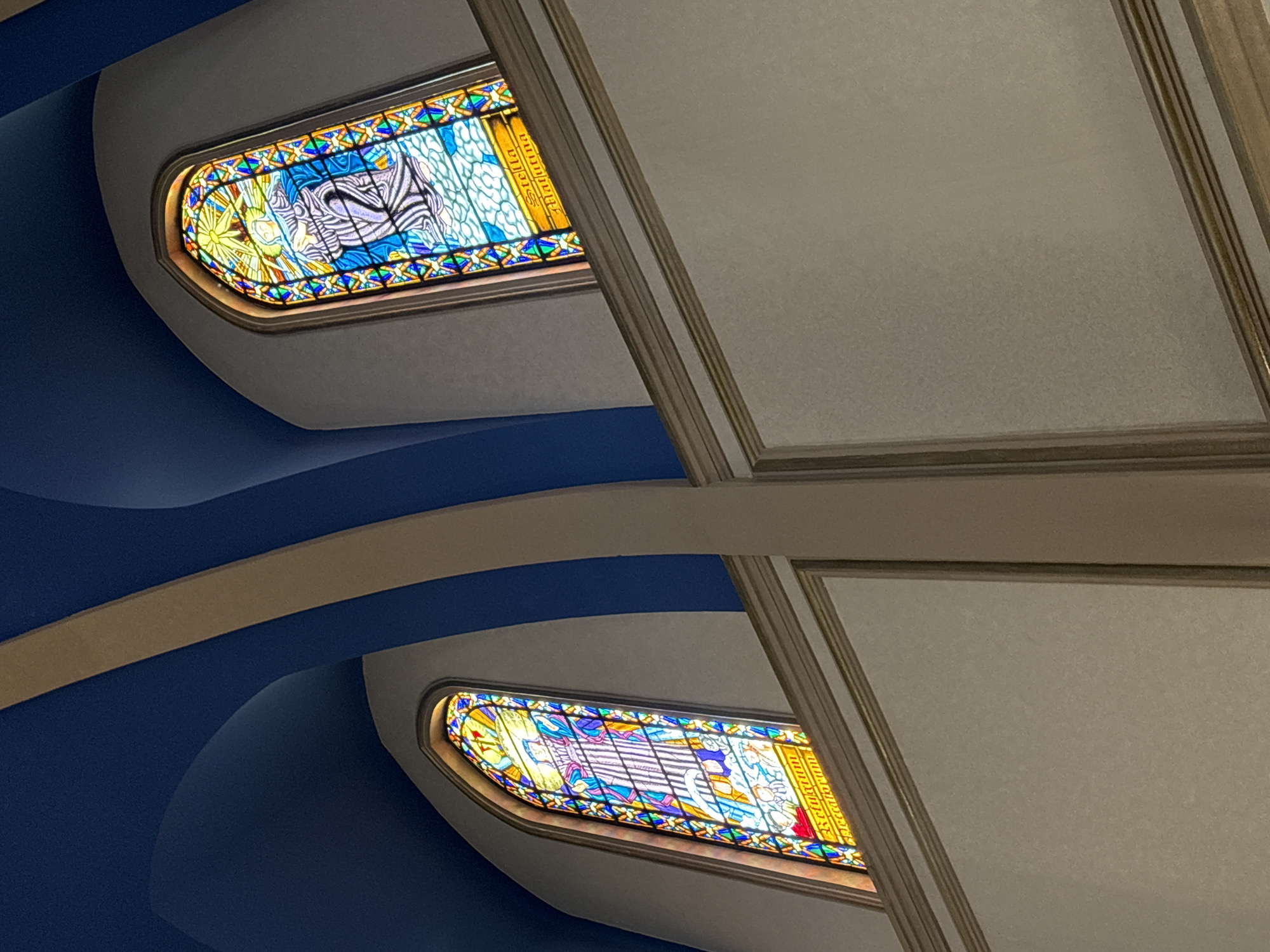 The image shows the interior of a church, specifically the ceiling and stained-glass windows. The ceiling is painted a deep blue and features curved architectural elements in a beige color. Two arched stained-glass windows are visible, one on the left and one on the right. The window on the left depicts a figure in colorful robes surrounded by geometric patterns and a sunburst. Below this figure, there are smaller figures and text in a language that appears to be Latin. The window on the right also features a figure in a white and purple robe with a halo and a sunburst. The lower portion of this window also contains text, which reads "Stella Matutina," and smaller figures. The walls are painted a neutral beige color and have decorative molding. The lighting suggests an indoor environment, possibly with light filtering through the stained glass. This image was taken inside the Metropolitan Cathedral of São Paulo, Brazil.