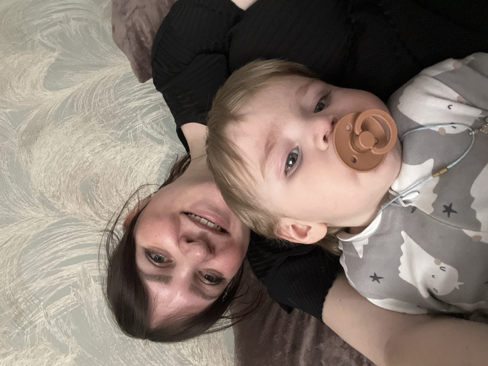 The image depicts an adult female and an infant in an indoor setting, reportedly in Noviy, Russia. The adult female, positioned behind and slightly to the left of the infant, has dark brown hair and is smiling directly at the camera. She wears a black ribbed top. The infant, with light brown hair and blue eyes, occupies the foreground, looking towards the camera with a terracotta-colored pacifier in its mouth. The infant wears a light grey long-sleeved garment patterned with white animals and black stars. A thin blue cord necklace with a cross-shaped pendant is visible on the infant's chest. Both individuals are supported by or resting against a soft, textured, brown-mauve colored surface, possibly bedding or a sofa. The background consists of a wall covered with light blue-grey wallpaper featuring a distinct white, feather-like, organic pattern.