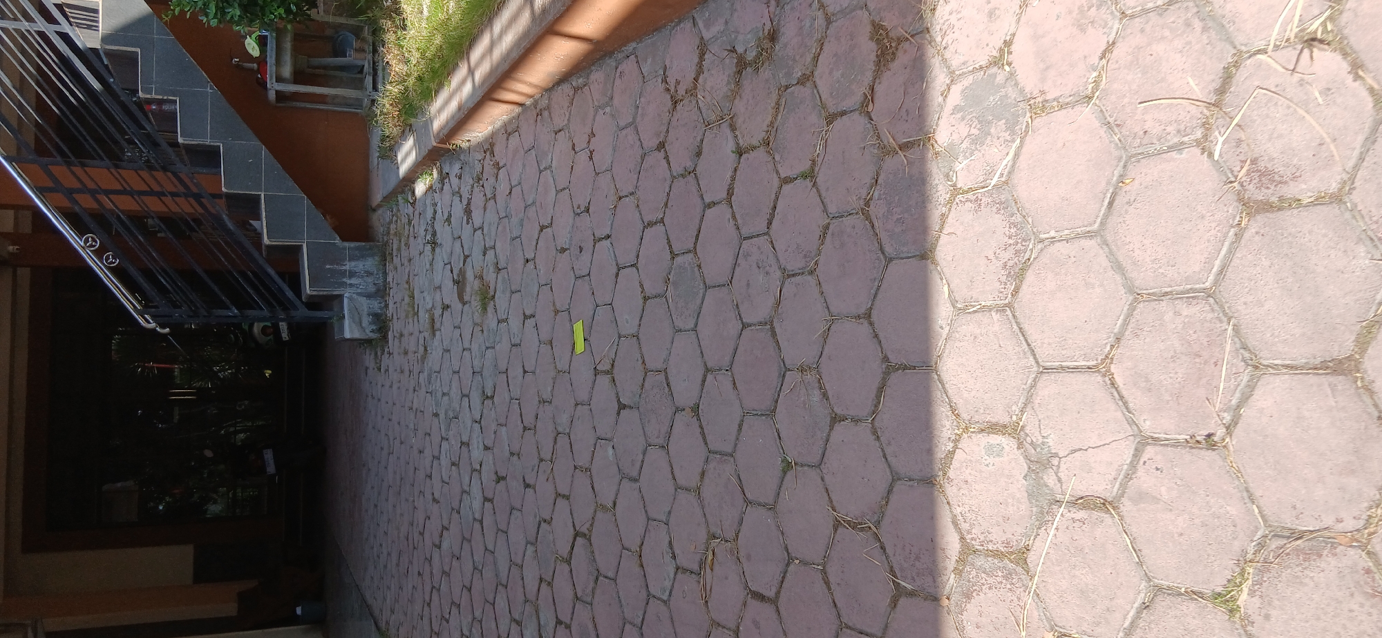 The image depicts a paved walkway made of hexagonal red bricks, located in front of a building in Pejaten, Indonesia. The walkway is partially in shadow, with sunlight illuminating the lower portion of the image. A small yellow object, possibly a piece of paper or a leaf, is visible on the bricks in the upper half of the walkway. To the right of the walkway, there is a raised concrete curb with grass growing along its edge, and beyond that, some vegetation. In the background, a staircase with a black metal railing leads up to an entrance of the building. The entrance appears dark, with some greenery visible inside. The scene is devoid of any people or apparent activity.