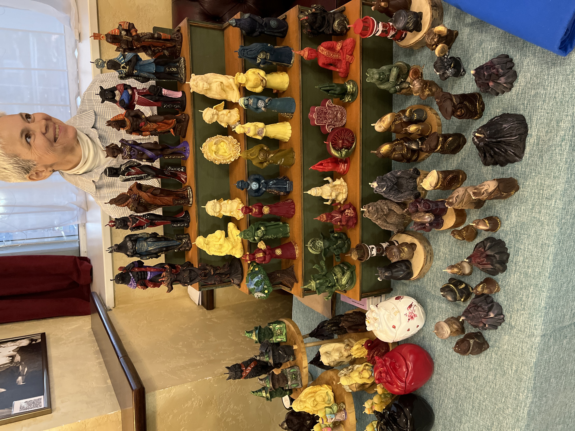 A person with short, white hair is partially visible on the left side of the frame. They are wearing a light-colored collared shirt and smiling.
The main focus of the image is a multi-tiered display stand showcasing a collection of figurines. The stand is made of wood and features multiple levels, each holding a variety of figurines that differ in color and design. The colors present in the figurines include browns, blacks, creams, yellows, reds, blues, and greens. Some figurines appear to be made of wood, while others may be made of other materials like resin or ceramic.
The figurines are arranged on a blue-green textured surface, possibly a tablecloth. Some of the figurines are placed on small wooden discs.
In the background, there is a window to the left of the person. Part of a dark-framed picture hanging on the wall and a dark-colored piece of furniture are visible in the lower-left corner of the frame.
There is no readily discernible information indicating the specific location in Petropavlovsk-Kamchatsky, Russia.