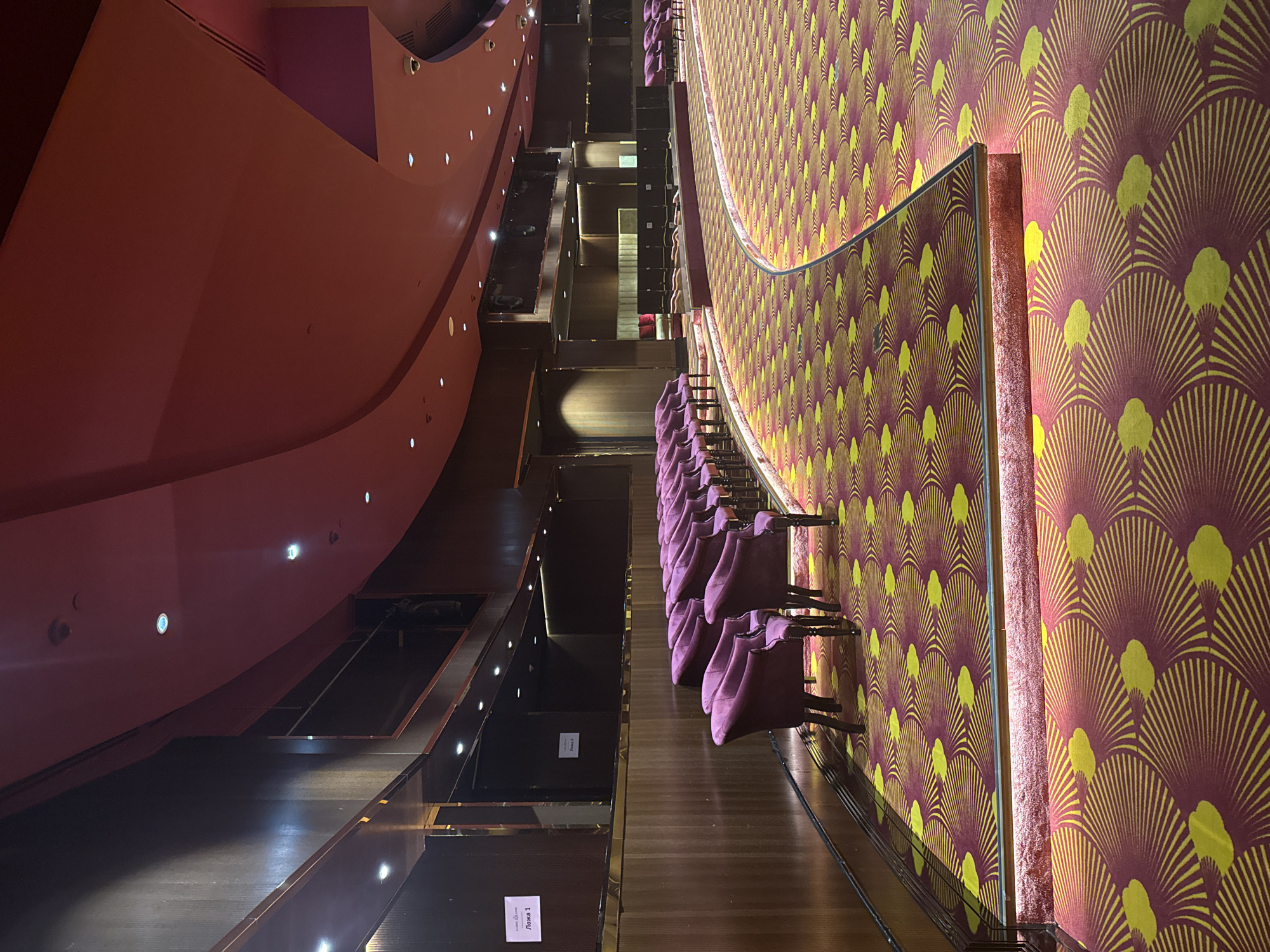 The image depicts an interior view of a theater or performance hall, likely in Estosadok, Russia. The scene is characterized by luxurious and ornate design elements. The dominant colors are deep reds, purples, and yellows, creating a rich and vibrant atmosphere.
The perspective is from a high vantage point, looking down onto a seating area. Rows of plush, deep purple upholstered chairs are arranged in curves, suggesting a sophisticated seating arrangement. These chairs are neatly placed on a floor that appears to be polished wood.
Adjacent to the seating area is a wall covered in a striking wallpaper featuring a repeating pattern of stylized fans or sunbursts in yellow and deep red or maroon. This bold design contributes to the opulent feel of the space.
To the left of the seating, a large, curved wall painted in a warm, rosy red dominates the scene. This wall has a smooth, matte finish and is punctuated by several small, circular lights embedded at intervals. The architecture of this wall suggests a grand, possibly semi-circular, structure common in performance venues.
The overall lighting appears to be dim, with the embedded lights on the red wall and a few other subtle light sources providing illumination. This suggests the image was taken either before a performance or during a period of low activity. There is no visible indication of a specific time of day or weather conditions, as the scene is entirely indoors.
There are a few pieces of visible text on small signs attached to some of the dark wooden panels in the lower left section of the image. One sign clearly reads "Ложа 1" (Lozha 1, meaning "Box 1" in Russian). Another sign above it appears to have similar Cyrillic text, possibly "Ложа 2" (Box 2). These signs indicate designated seating areas, further confirming the setting as a venue with tiered or boxed seating.
The image captures a sense of elegance and anticipation, characteristic of a high-end entertainment space. No people are visible in the frame, focusing the viewer's attention solely on the architectural and decorative elements of the venue.