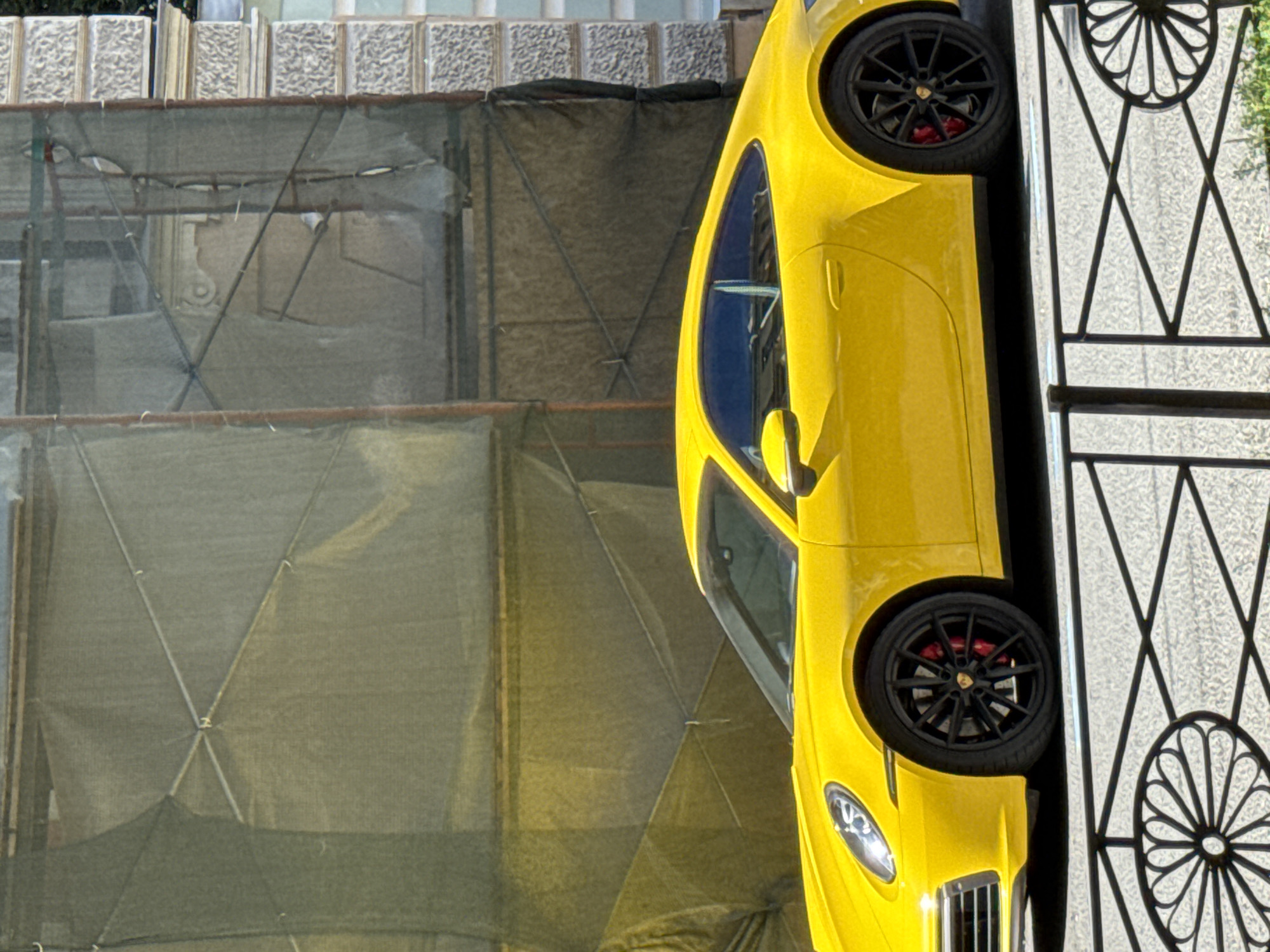 The media file displays a close-up, vertically oriented shot of a bright yellow sports car, likely a Porsche, parked next to a decorative black metal fence. The car's sleek design is highlighted, with a portion of its front fender, headlight, wheel, and side profile visible. The wheel features black spokes and red brake calipers.
The car appears to be parked on a street, as indicated by the textured pavement visible through the fence. Behind the car, there's a building with decorative stone elements, possibly an apartment building or a historical structure. In the background, scaffolding covered with green netting suggests ongoing construction or renovation work.
The lighting indicates it is daytime, with sunlight casting shadows. The scene appears to be set in an urban environment, with architectural features and construction materials suggesting a city location. There is no visible text within the frame. The overall impression is a snapshot of a luxury car in an everyday urban setting that is undergoing some form of development.