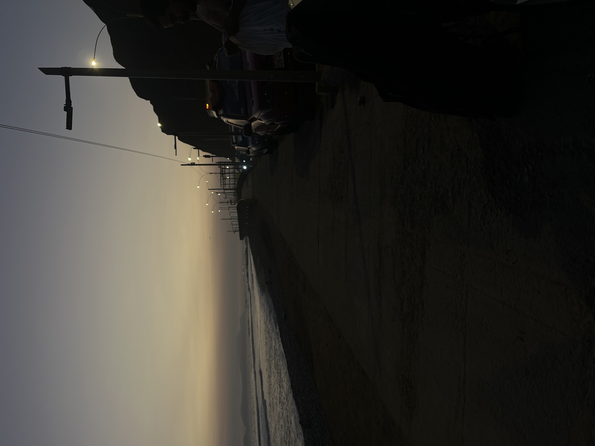 The image depicts a dusky scene along a beach promenade in Miraflores, Peru. The sky is a gradient of pale yellow and gray, indicating twilight. To the left, the ocean is visible with gentle waves lapping against a pebble beach. A concrete walkway lines the beach, transitioning into a darker, sandy area in the foreground.
Along the promenade, several vehicles are parked, including what appears to be a red SUV with a license plate ending in "C2G-43", and other dark-colored cars. Streetlights with a warm glow illuminate the walkway, casting long shadows. In the background, a dark, rocky hillside forms a dramatic backdrop to the scene.
On the right side of the image, a person is partially visible, silhouetted against the dim light. They are seen from the chest up, with their arms and hands engaged in an action, possibly handling a white object or fabric. The person appears to be in a seated or hunched position. The overall atmosphere is quiet and serene, capturing a moment at the end of the day in this coastal district of Lima.