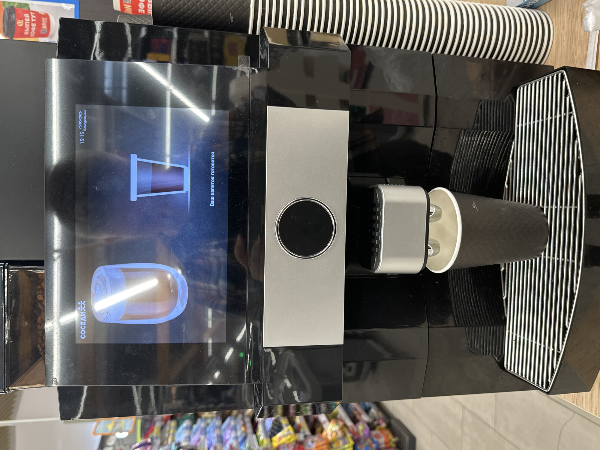 The image depicts a coffee machine in what appears to be a store, likely in Бараўлянскі сельскі Савет, Belarus. The coffee machine is black and silver, with a digital display showing the process of coffee preparation. A stack of paper coffee cups is next to the machine, and a coffee cup is being filled by the machine. In the background, there are shelves of various products, suggesting a retail environment. The time on the screen is 15:15 on Monday the 25th of May 2026. The machine's screen reads "Your drink is being prepared." A logo for "Neighbors" is on the screen.