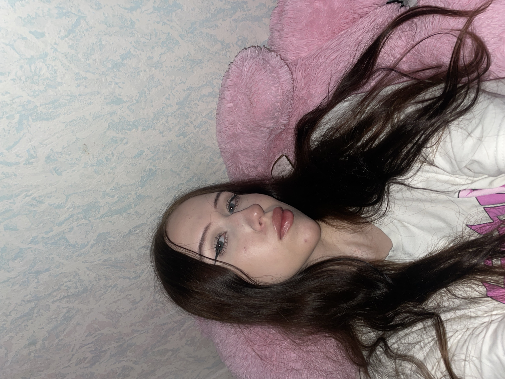 In Votkinsk, Russia, a young woman with long, dark brown hair is pictured, looking directly at the camera. She is wearing a white t-shirt with a pink graphic design. Her makeup includes eyeliner and lip gloss. She is leaning against a large, fuzzy pink object, possibly a stuffed animal or cushion. The background features a wall with a textured, light blue and white pattern.