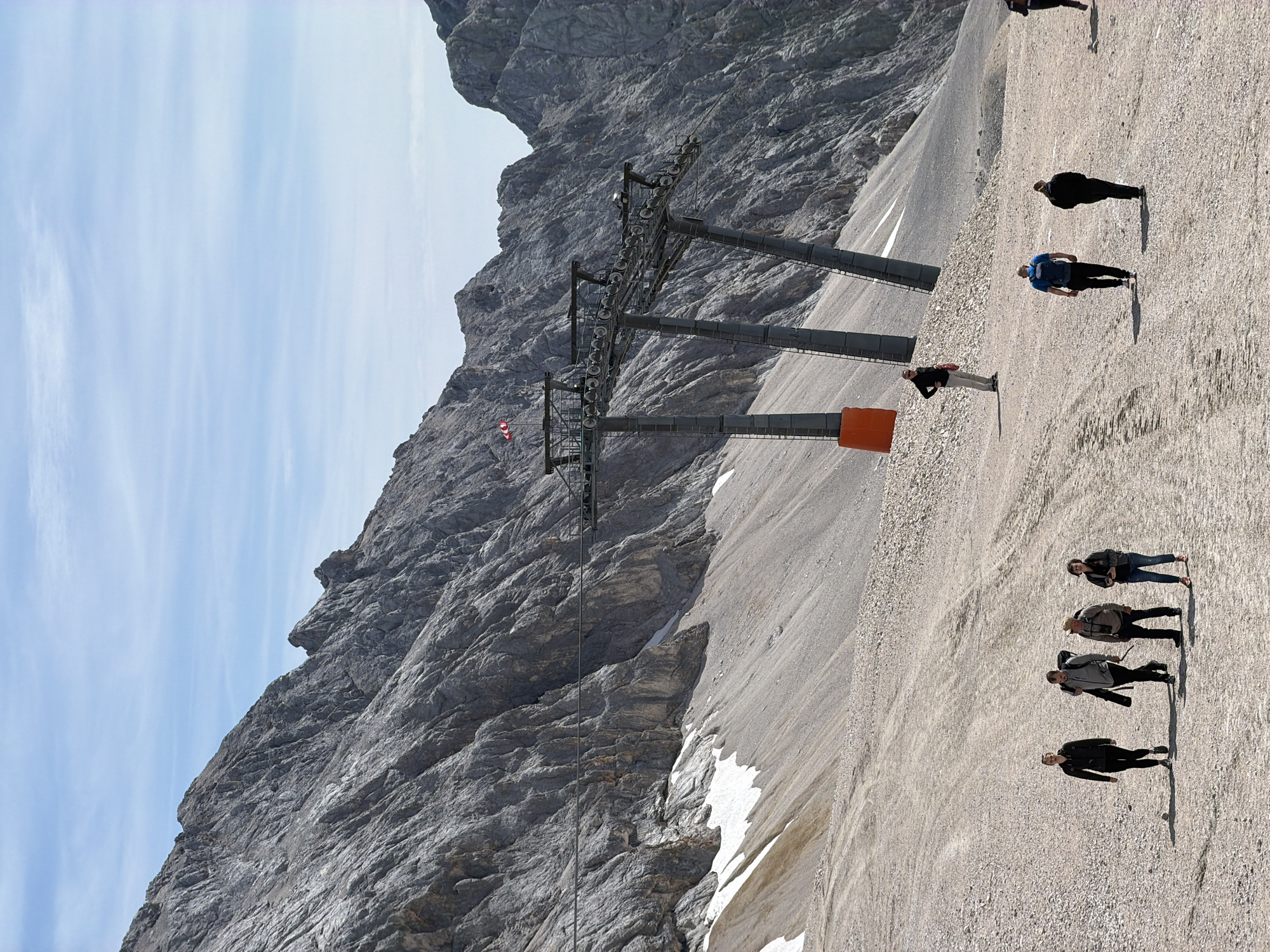 In Wildermieming, Austria, a group of people walk on a gravelly mountain slope under a bright, partly cloudy sky. In the background, a large ski lift structure with multiple towers and numerous pulleys stands against a backdrop of rugged, grey mountains. Patches of snow are visible on the mountainside below the ski lift. The people are scattered across the slope, some walking individually and others in small groups. The terrain is uneven, covered in small stones and dirt, with tire tracks visible in places, suggesting vehicle or machinery use. The overall scene depicts a mountainous recreational area, possibly a ski resort in its off-season, with the imposing presence of the ski lift and the natural grandeur of the Alps.